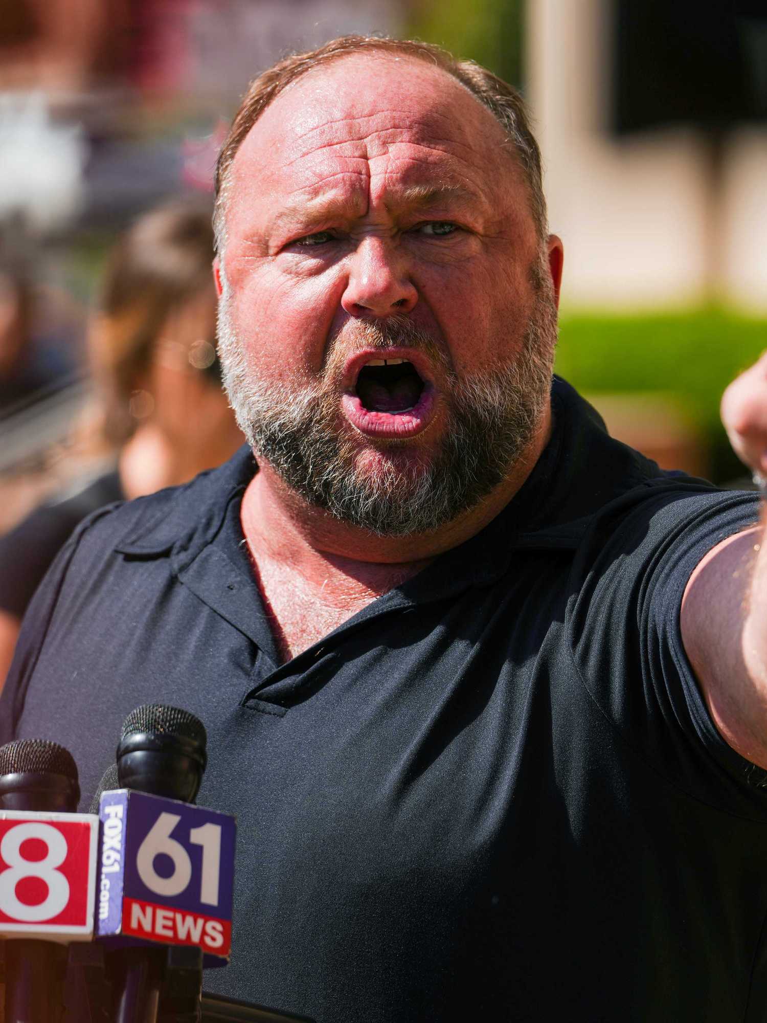 Alex Jones, a bearded white man with grey hair and a black shirt, points and shouts angrily at someone off-camera to his left.