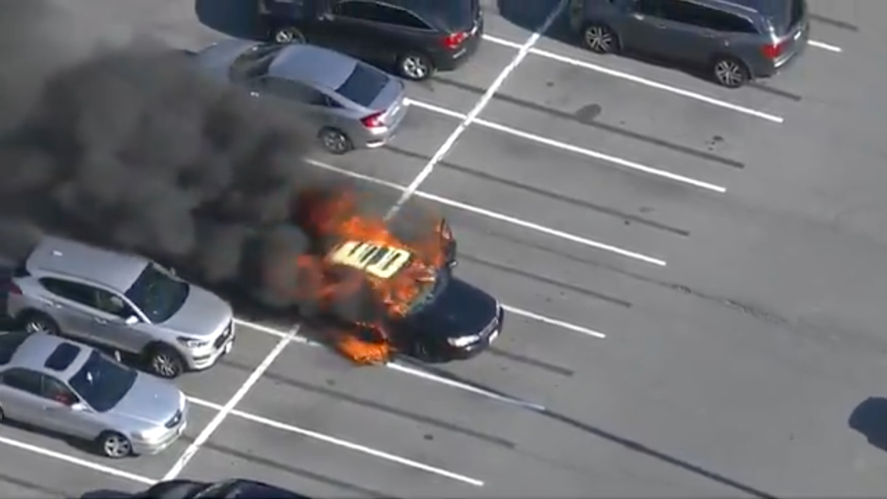 An aerial shot of a black car in flames in the middle of a car park.