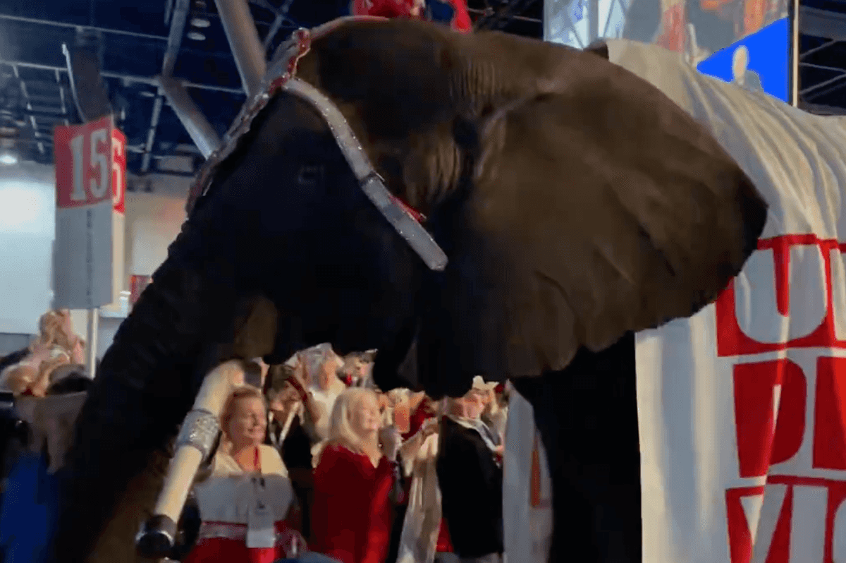 An elephant walking through a convention hall, with a banner draped over its back reading: 'Unity drives victory'.
