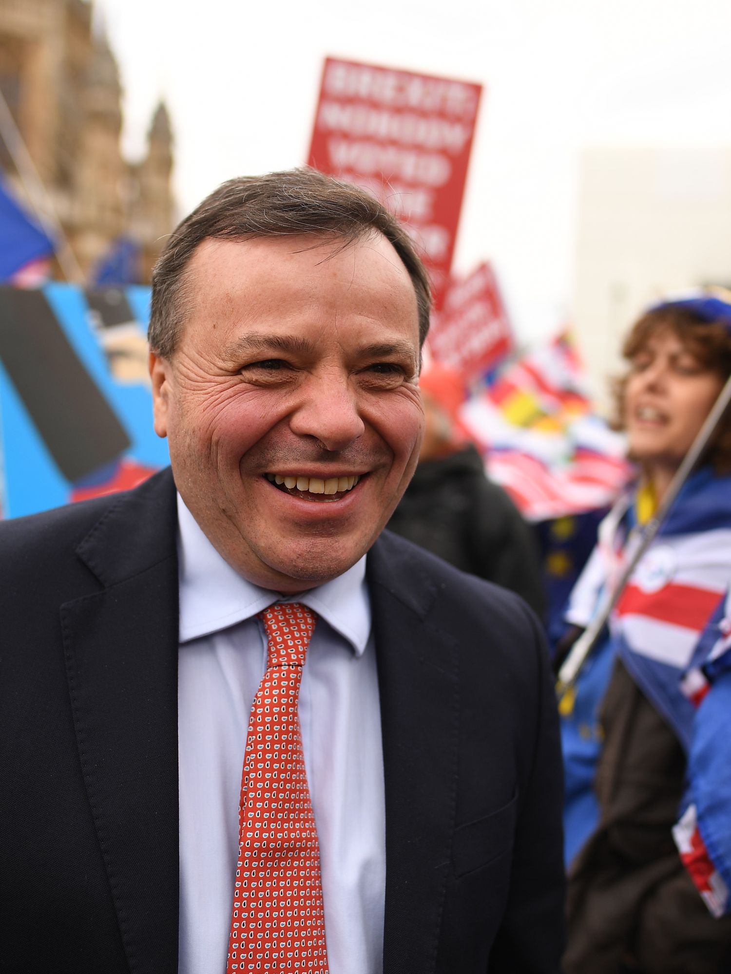 Arron Banks, a white man with short black hair in a black suit, smiles as anti-Brexit protesters hold up placards behind him.