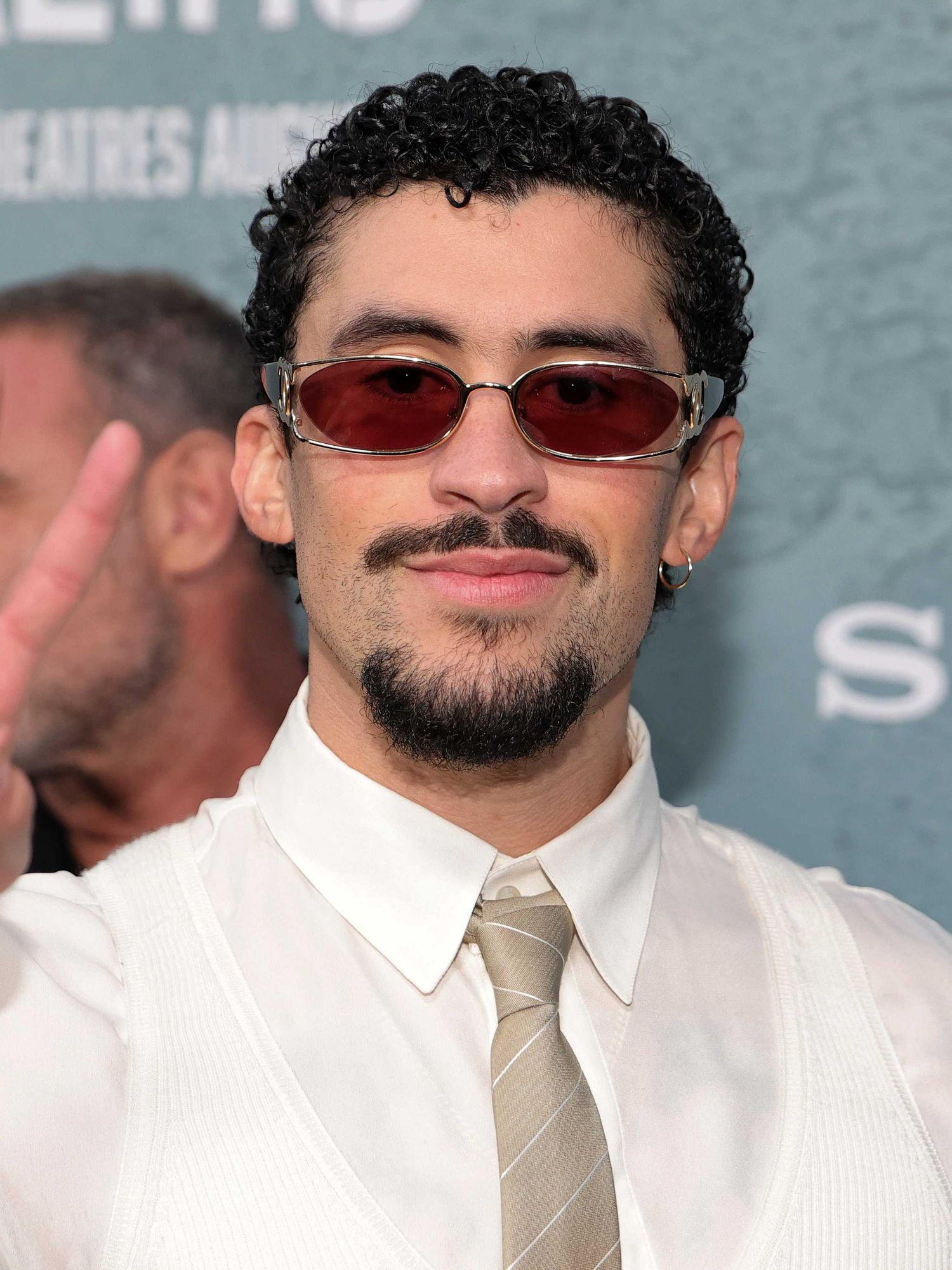 Bad Bunny, a Puerto Rican man with short curly black hair, a moustache and beard, smiles at the camera while holding up a peace sign.