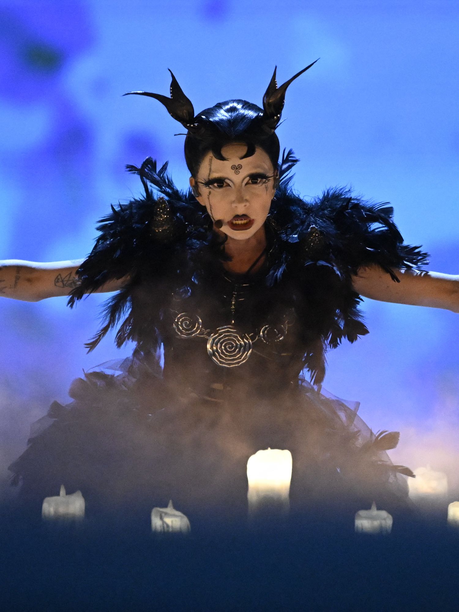 Bambie Thug, a white person in a black feathered dress and a horned headwear, has their arms outstretched to the side as they perform during Eurovision.