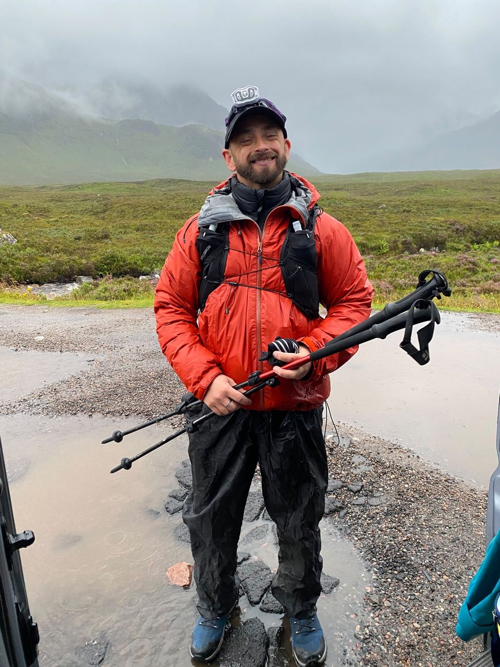 Benjamin Ward smiling, standing in a rural area wearing walking gear and holding walking poles