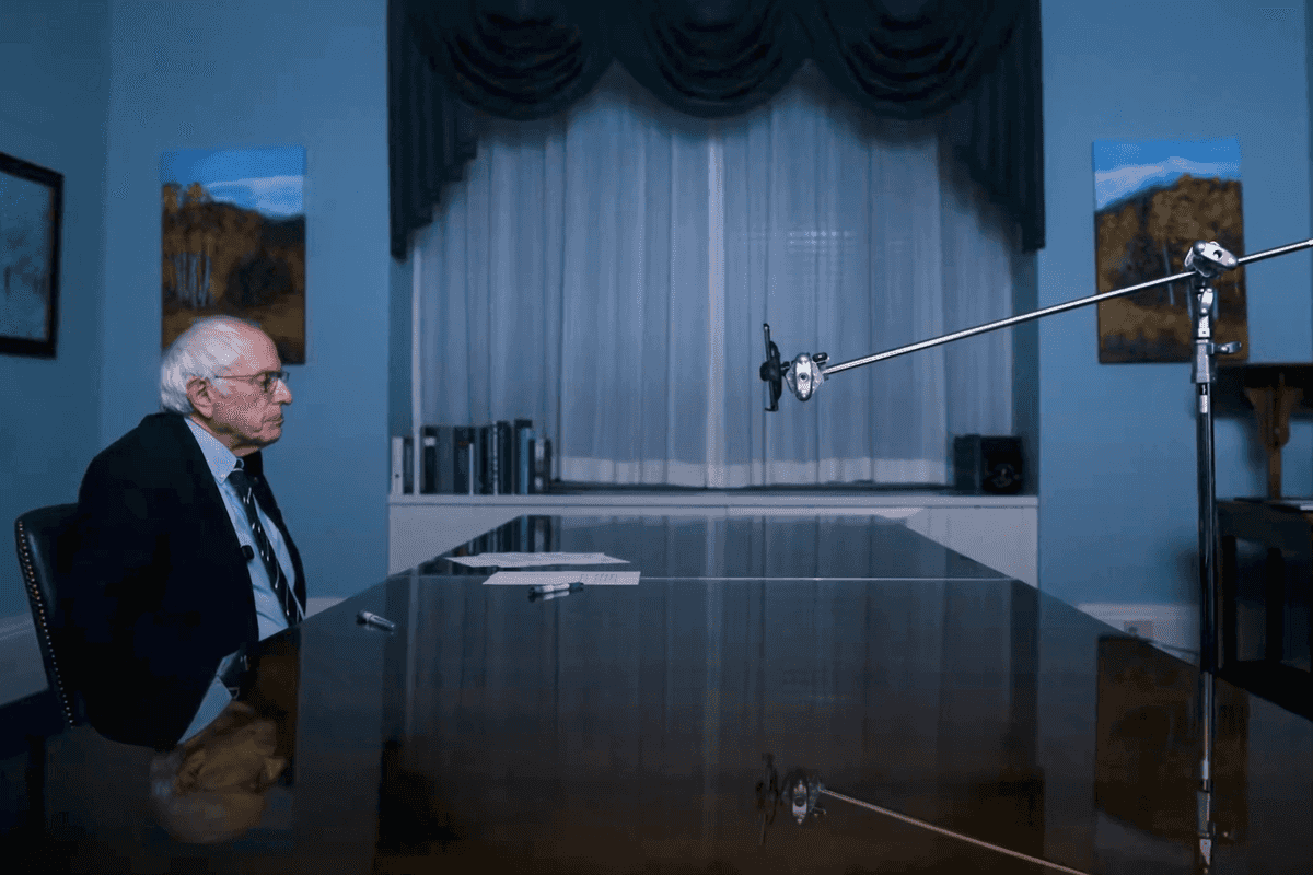 Bernie Sanders, left, a white man with glasses, short white hair and a suit, sits at a table opposite an iPhone on a stand.