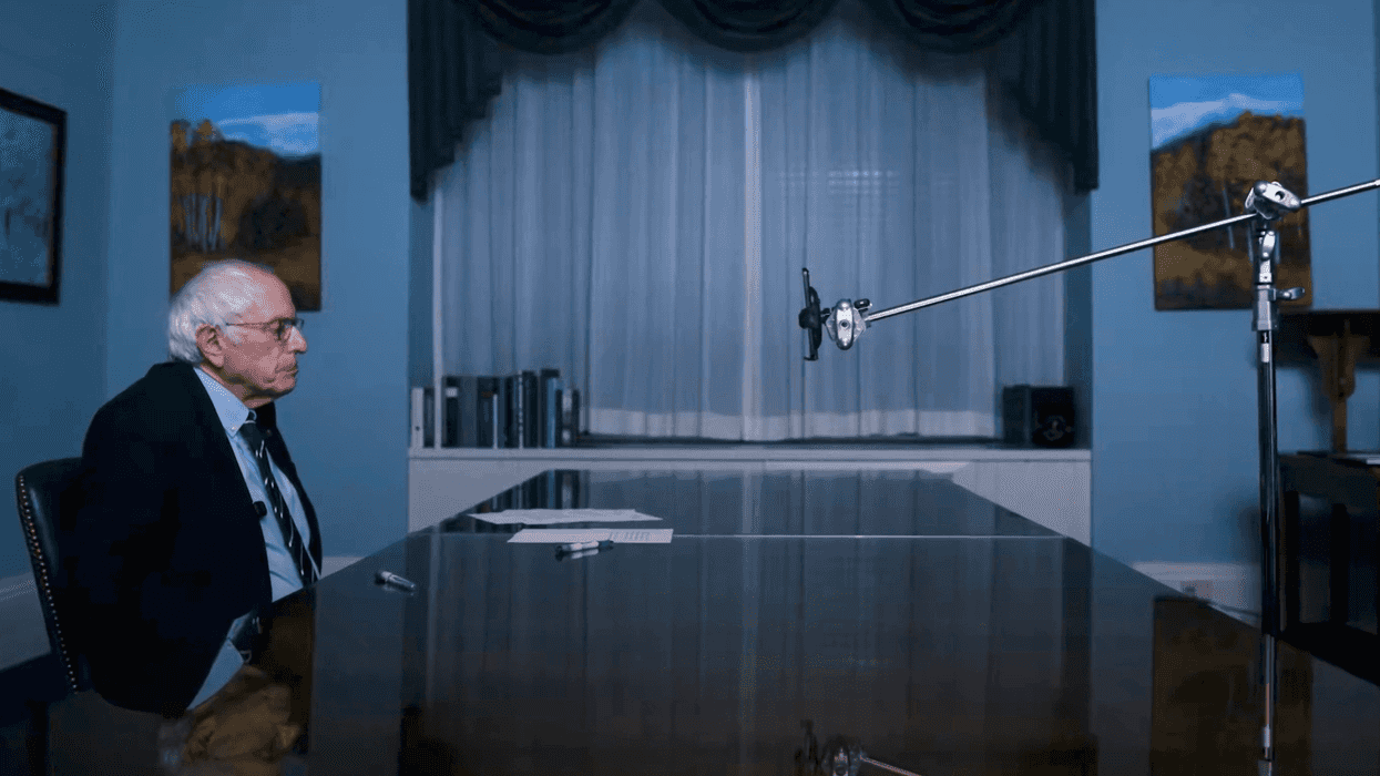 Bernie Sanders, left, a white man with glasses, short white hair and a suit, sits at a table opposite an iPhone on a stand.