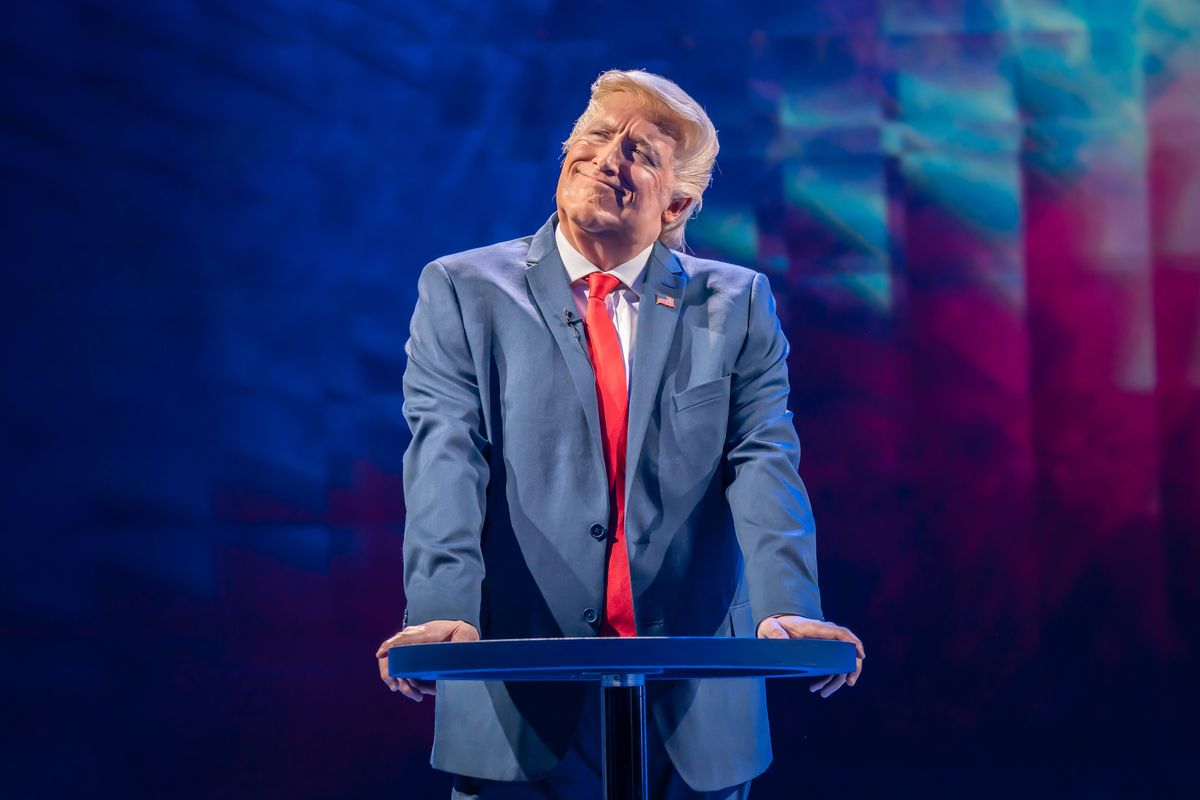 Bertie Carvel as Donald Trump, a white man with short blonde hair, a blue suit and red tie, resting his hands on a lectern. He's tilting his head slightly and has a smug smile on his face.