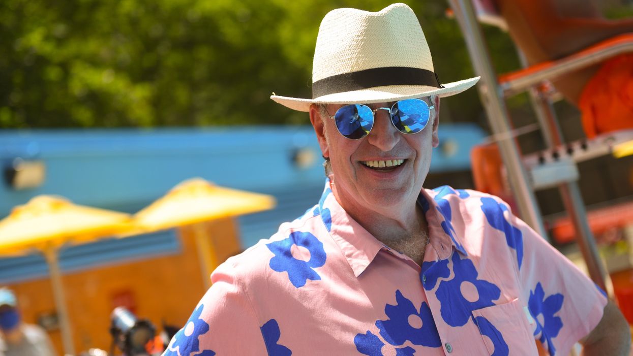 Bill de Blasio, a white man, is at a pool, smiling and wears a white hat, pink floral shirt with blue flowers, and blue tinted sunglasses, which reflect a woman’s chest.