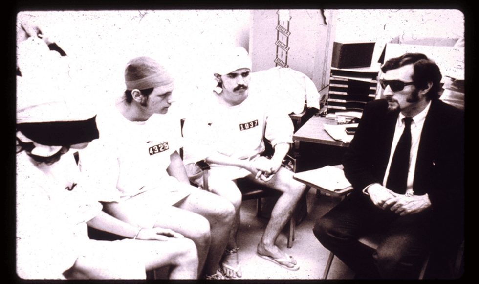 Black and white photo. Male participants in the Stanford Prison Experiment, in white numbered T-shirts, sit opposite a man in a black suit and tie and sunglasses.
