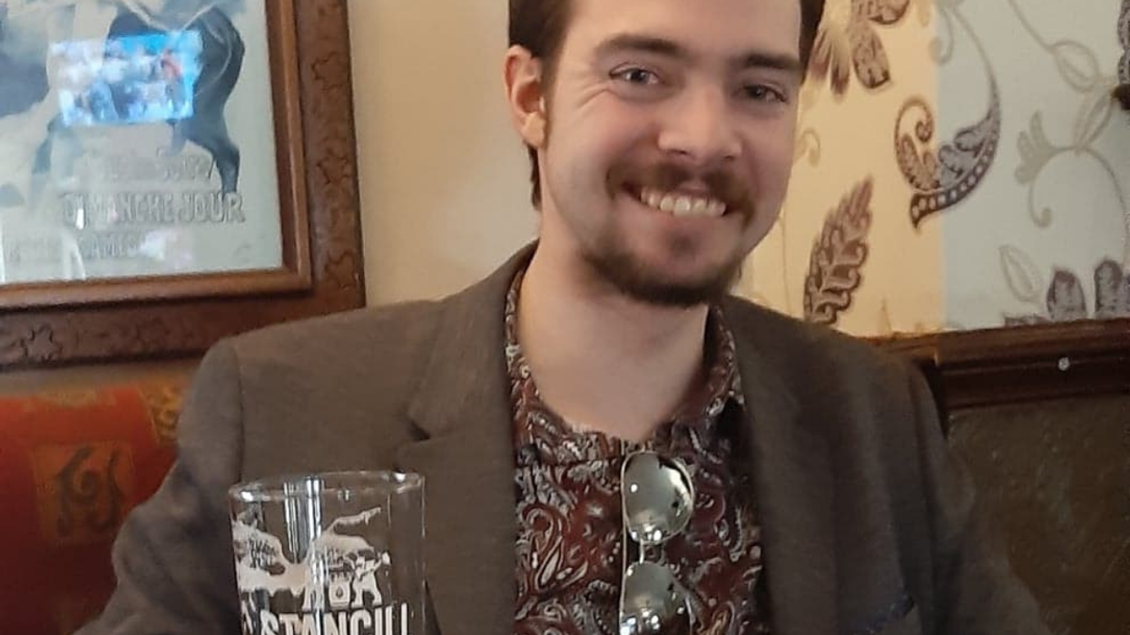 Bob, a white man with a moustache and beard, wears a red patterned shirt and brown coat. He is smiling, and holds a pint of beer in his right hand. He is in a pub.