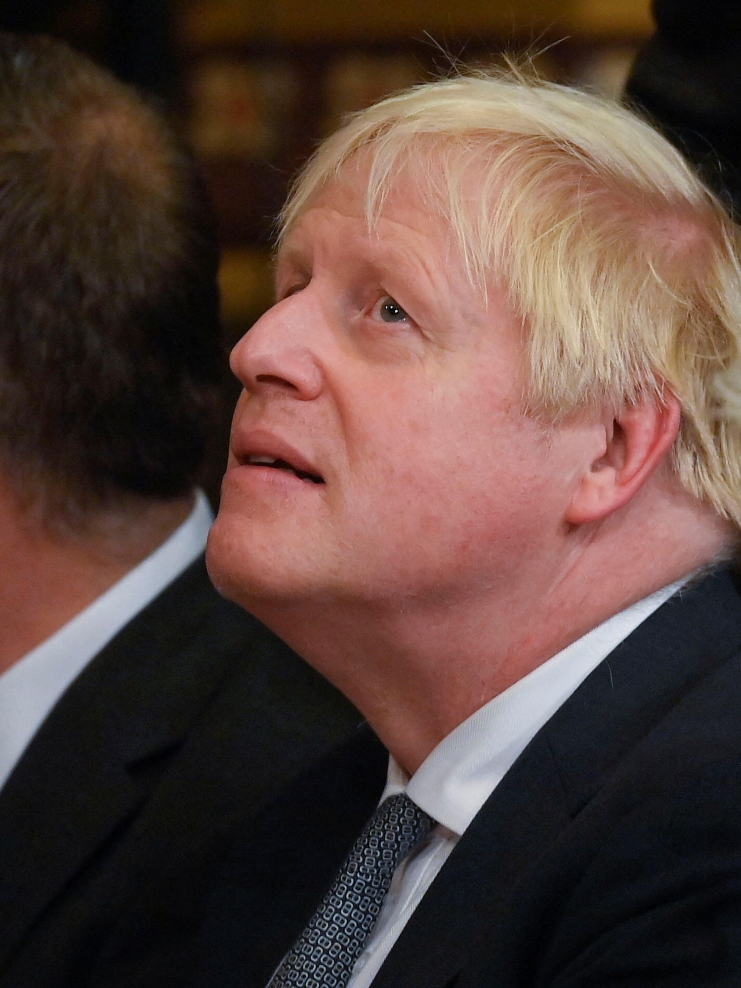 Boris Johnson, a white man with bushy blonde hair, tilts his head slightly upwards in concen. A white man is sat to his right and a white woman to his left.