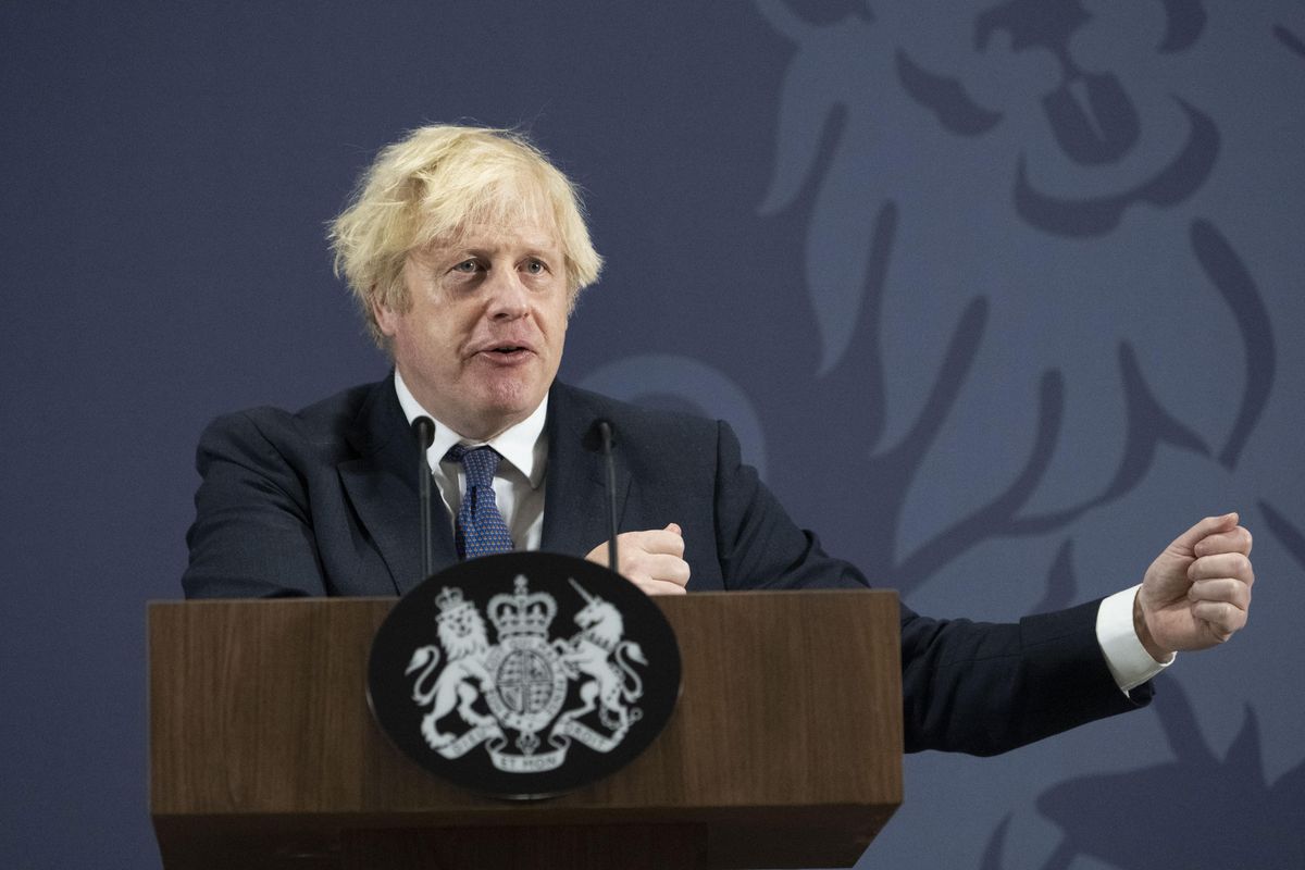 Boris Johnson, a white man with scruffy blonde hair and a black suit, gestures with his hands behind a wooden lectern.