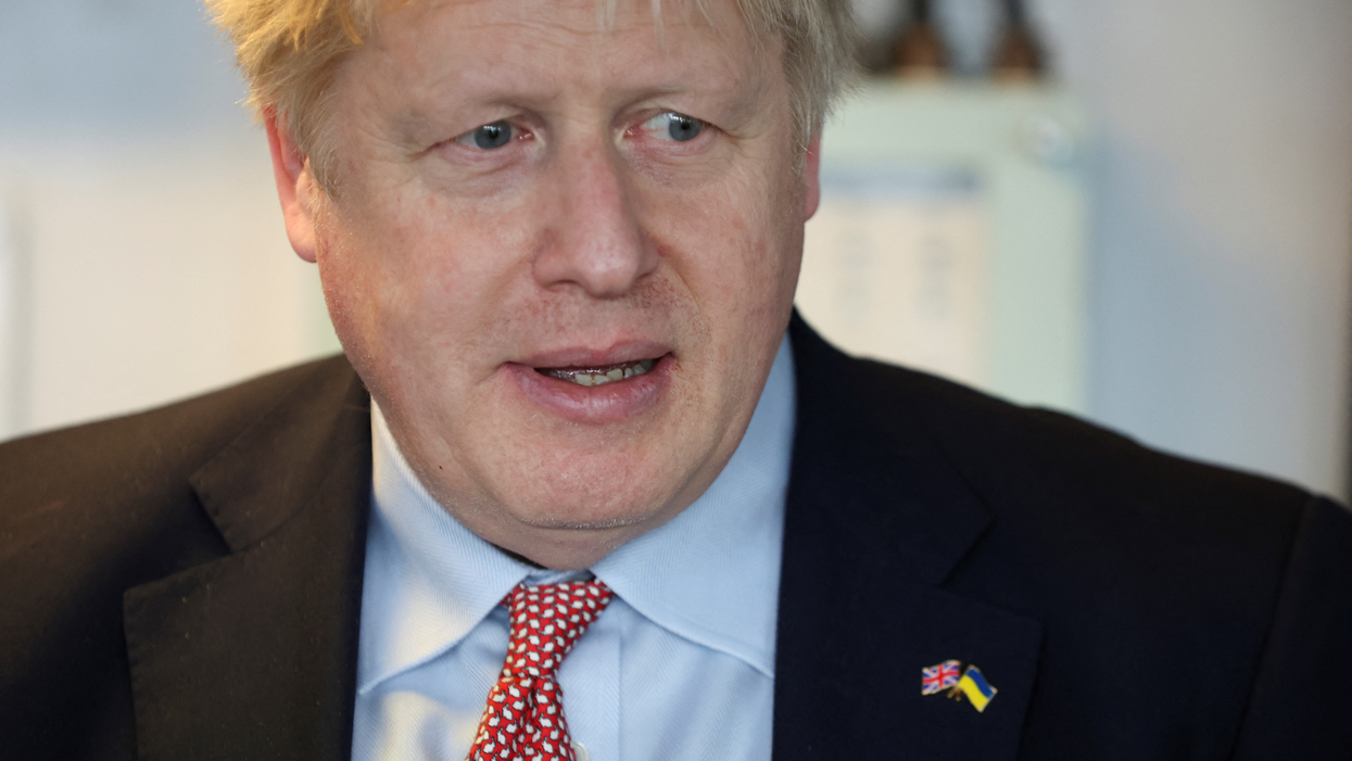 Boris Johnson, a white man with scruffy blonde hair, wears a suit and is looking downwards, slightly to the left. On his black suit jacket is a pin showing the UK flag and the Ukrainian flag together.