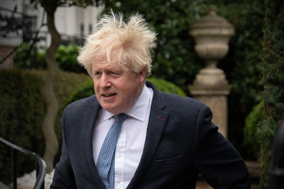 Boris Johnson, a white man with short blonde hair blowing in the wind, wearing a black suit and walking down a street.