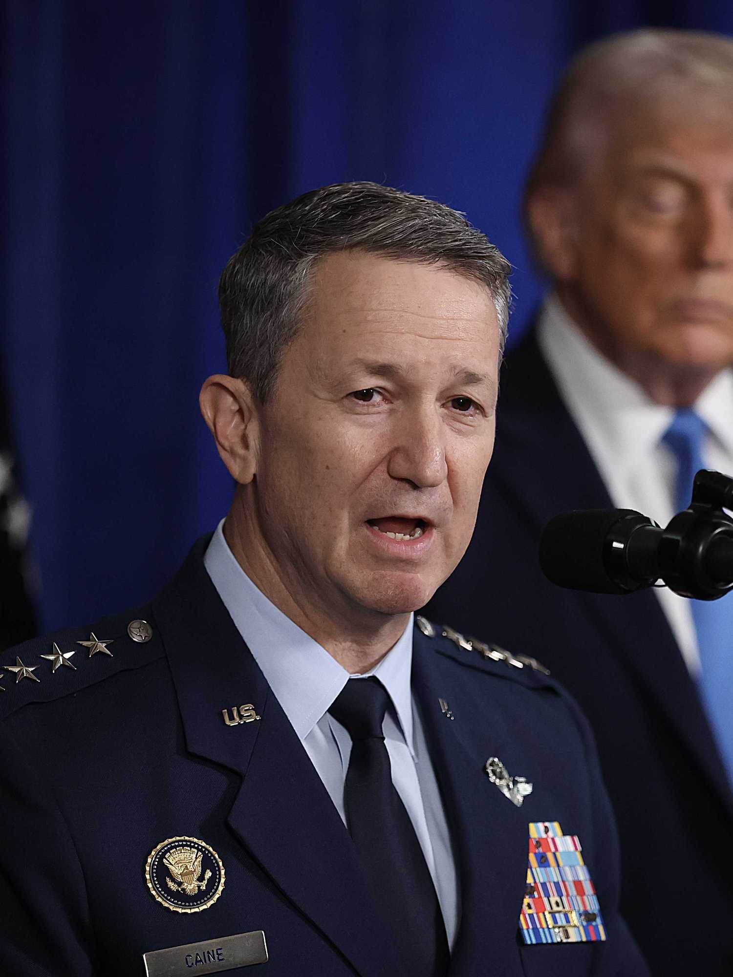 Chairman of the Joint Chiefs of Staff Dan Caine speaking during a press conference, with Donald Trump standing out of focus in the background with his eyes closed.