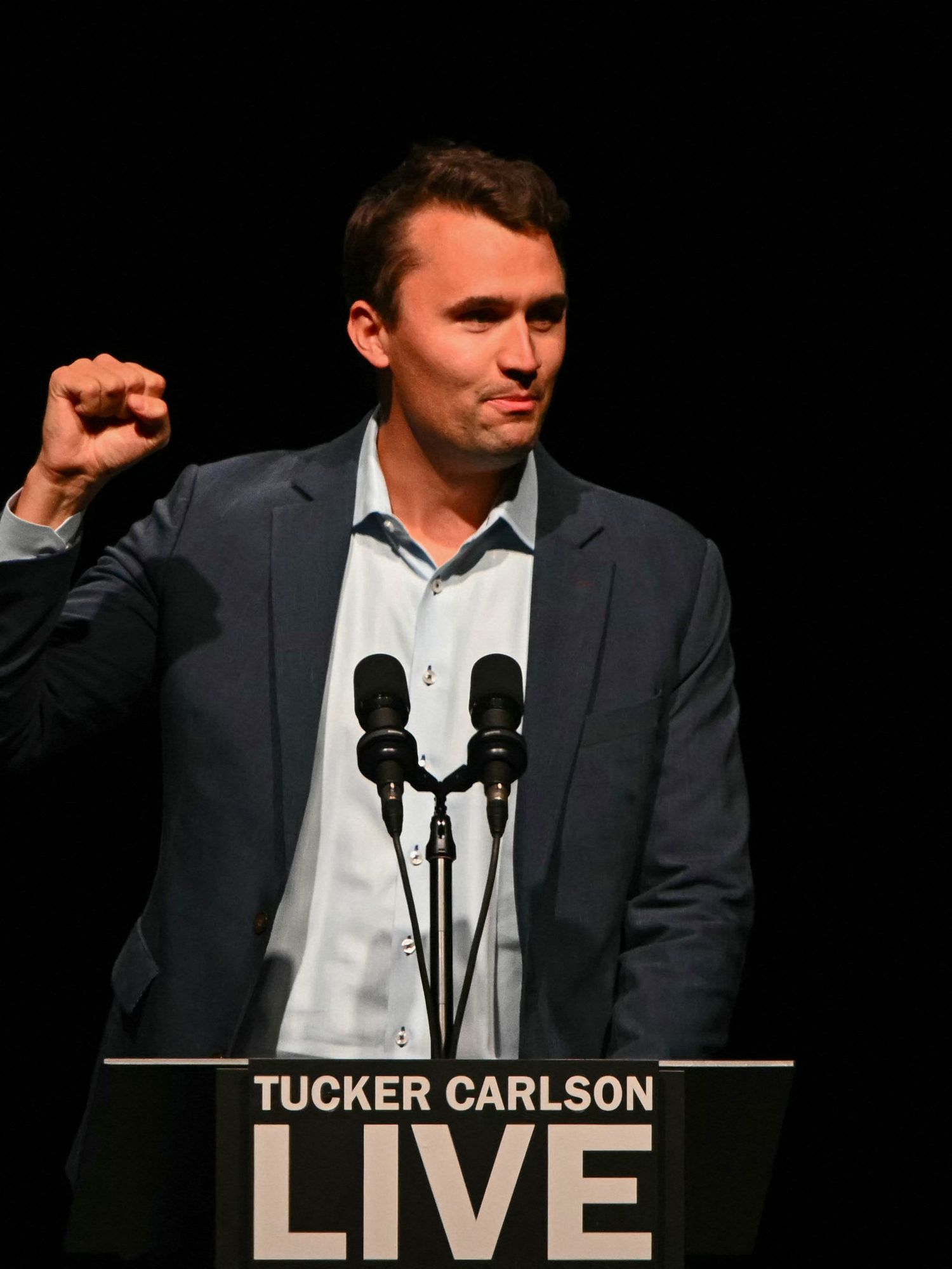 Charlie Kirk raises his right fist, smiling, as he stands behind a lectern with 'Tucker Carlson Live' on it.