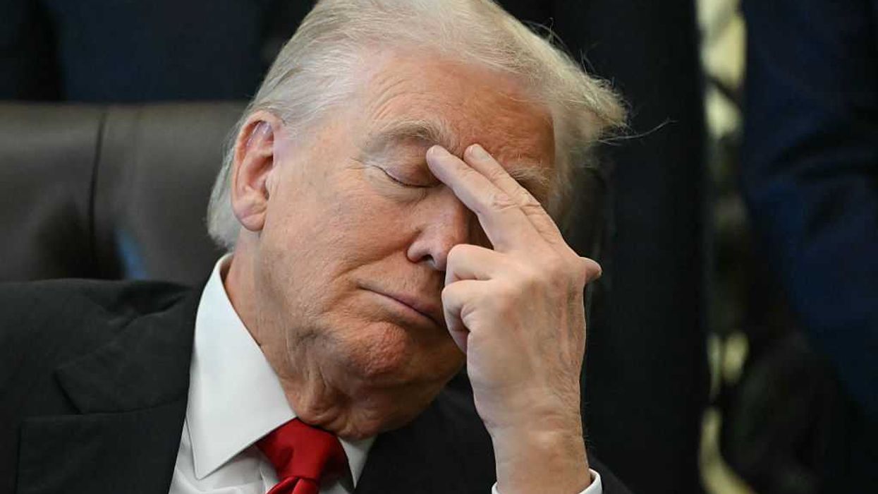Close-up image of Donald Trump, with his eyes closed, placing his left index finger and middle finger to his right eyebrow as he sits in his chair in the Oval Office.