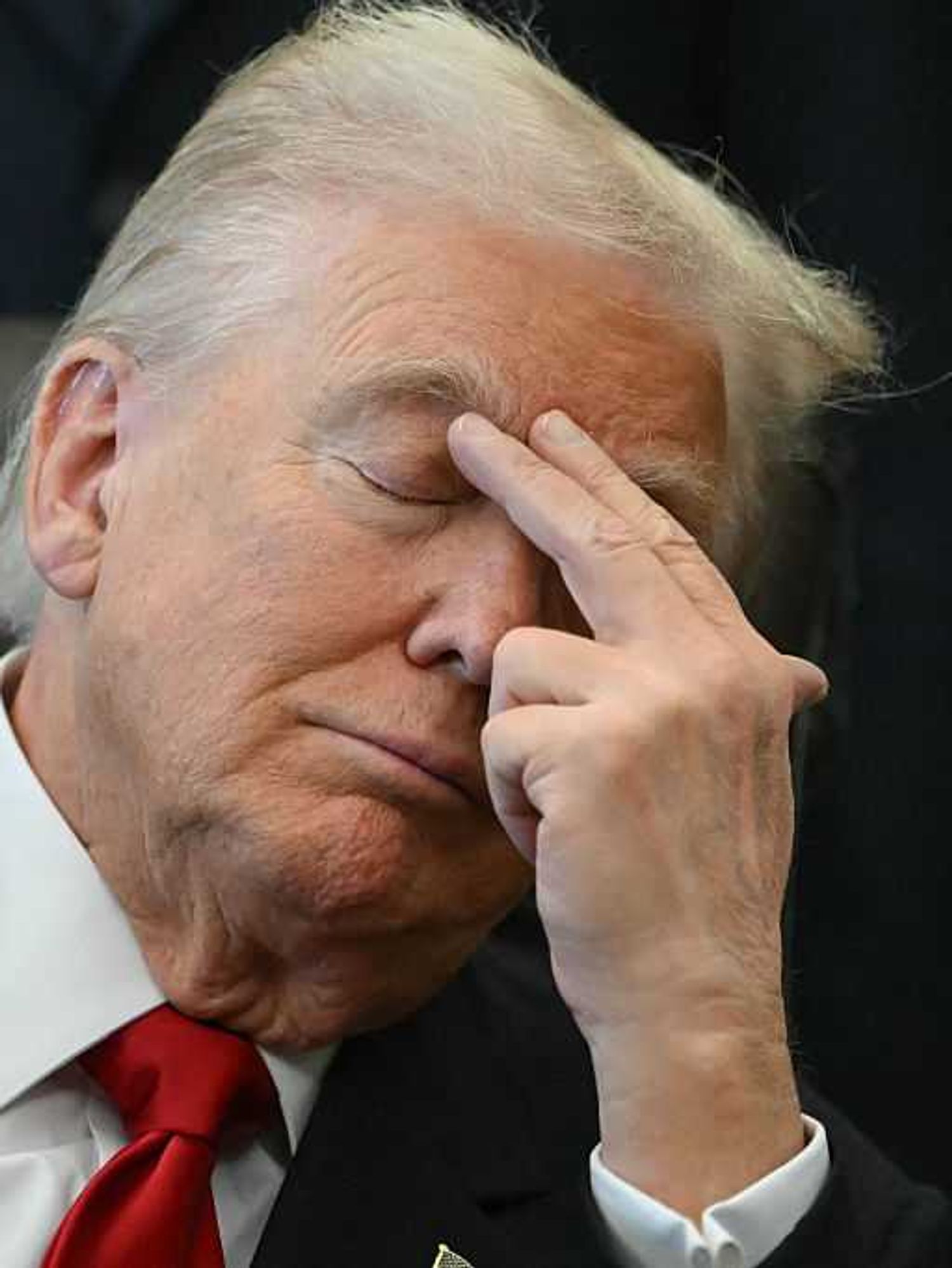 Close-up image of Donald Trump, with his eyes closed, placing his left index finger and middle finger to his right eyebrow as he sits in his chair in the Oval Office.