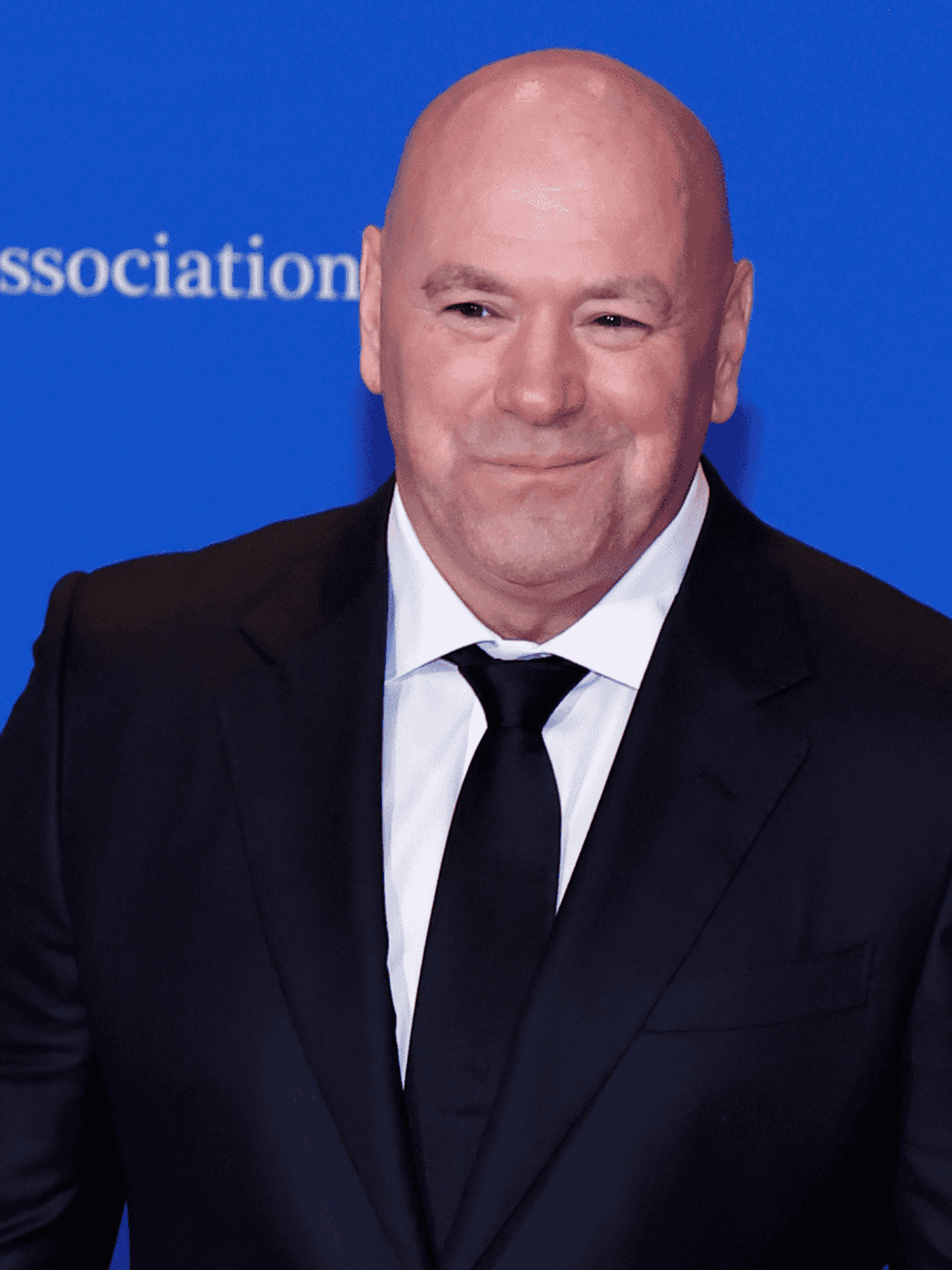Dana White, a bald white man in a black suit and tie, smiles in front of a blue poster board with the text 'White House Correspondents Association' on it.