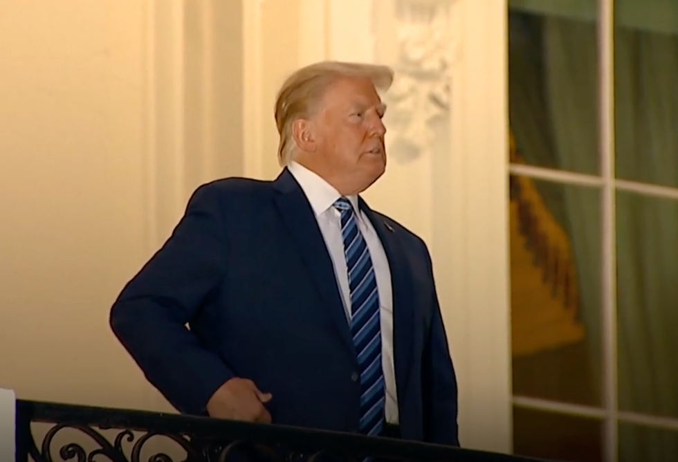 Donald Trump, a blonde white man in a navy suit, stands on a balcony at The White House and has his right hand in his right suit pocket.