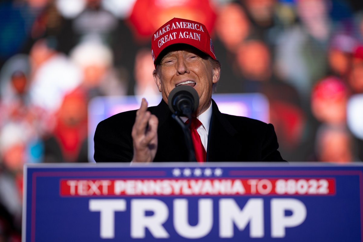 Donald Trump, a white man with a red Make America Great Again cap, speaking at a rally behind a lectern with a blue Trump sign on it.