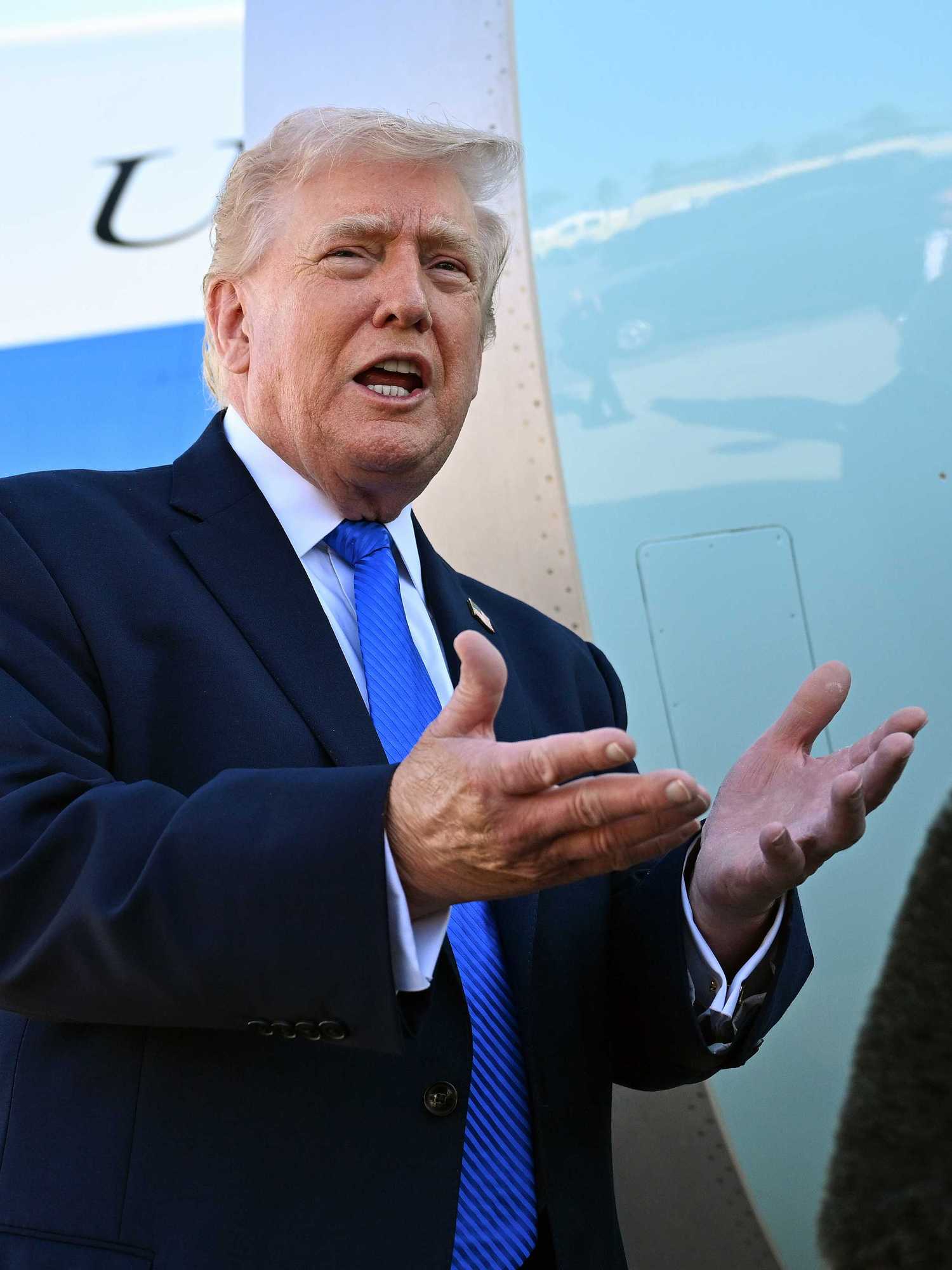Donald Trump, a white man with short blonde hair and a black suit, talking to reporters next to Air Force One.