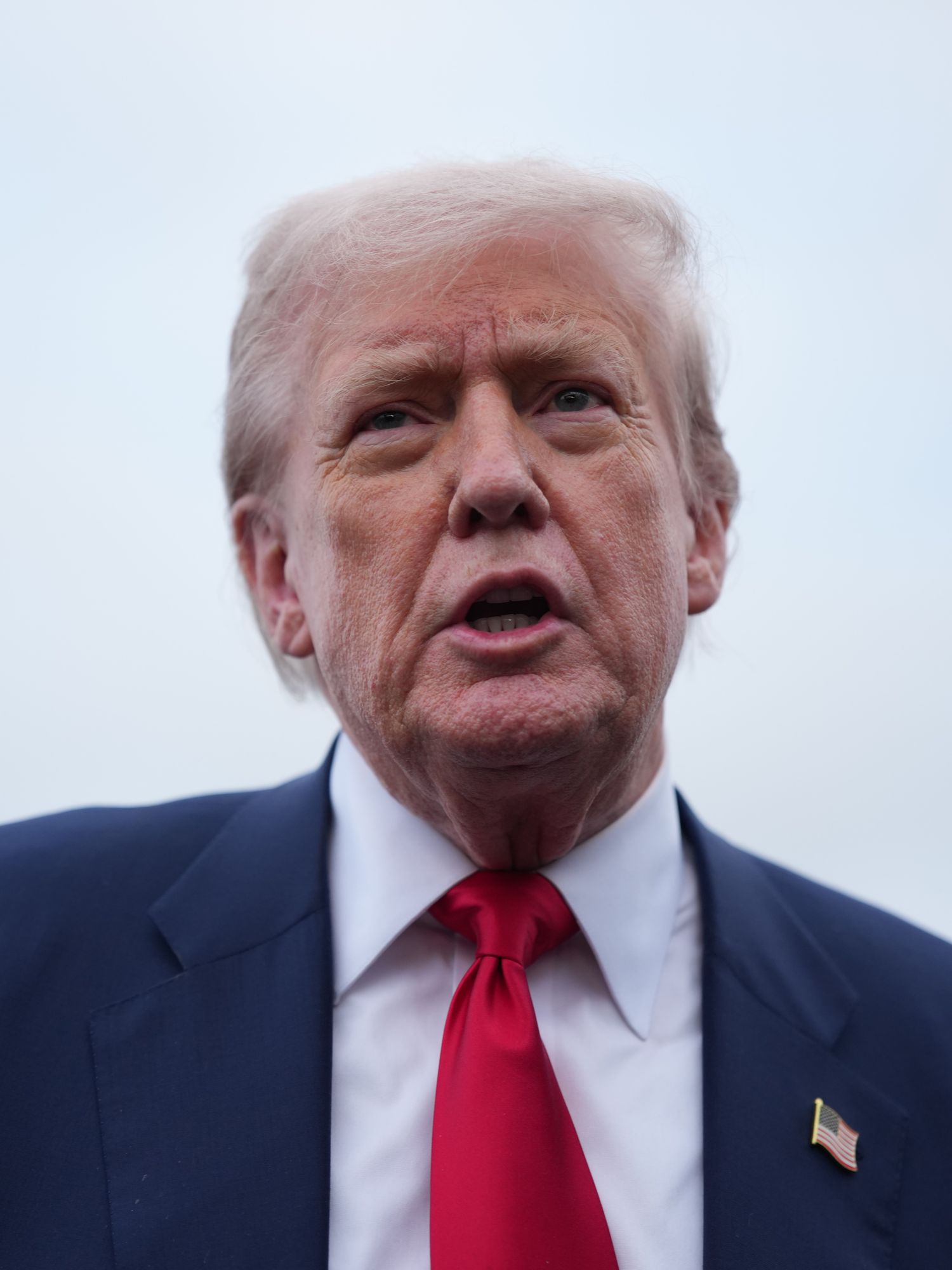 Donald Trump, a white man with short blonde hair and a blue suit and red tie, with grey skies behind him.