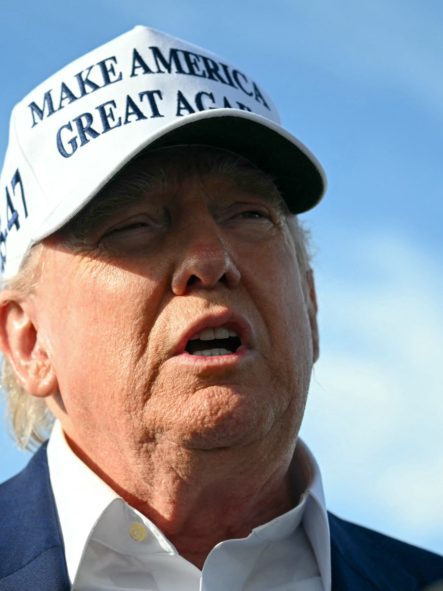 Donald Trump, a white man with short blonde hair and a white MAGA cap, talks to reporters outside.