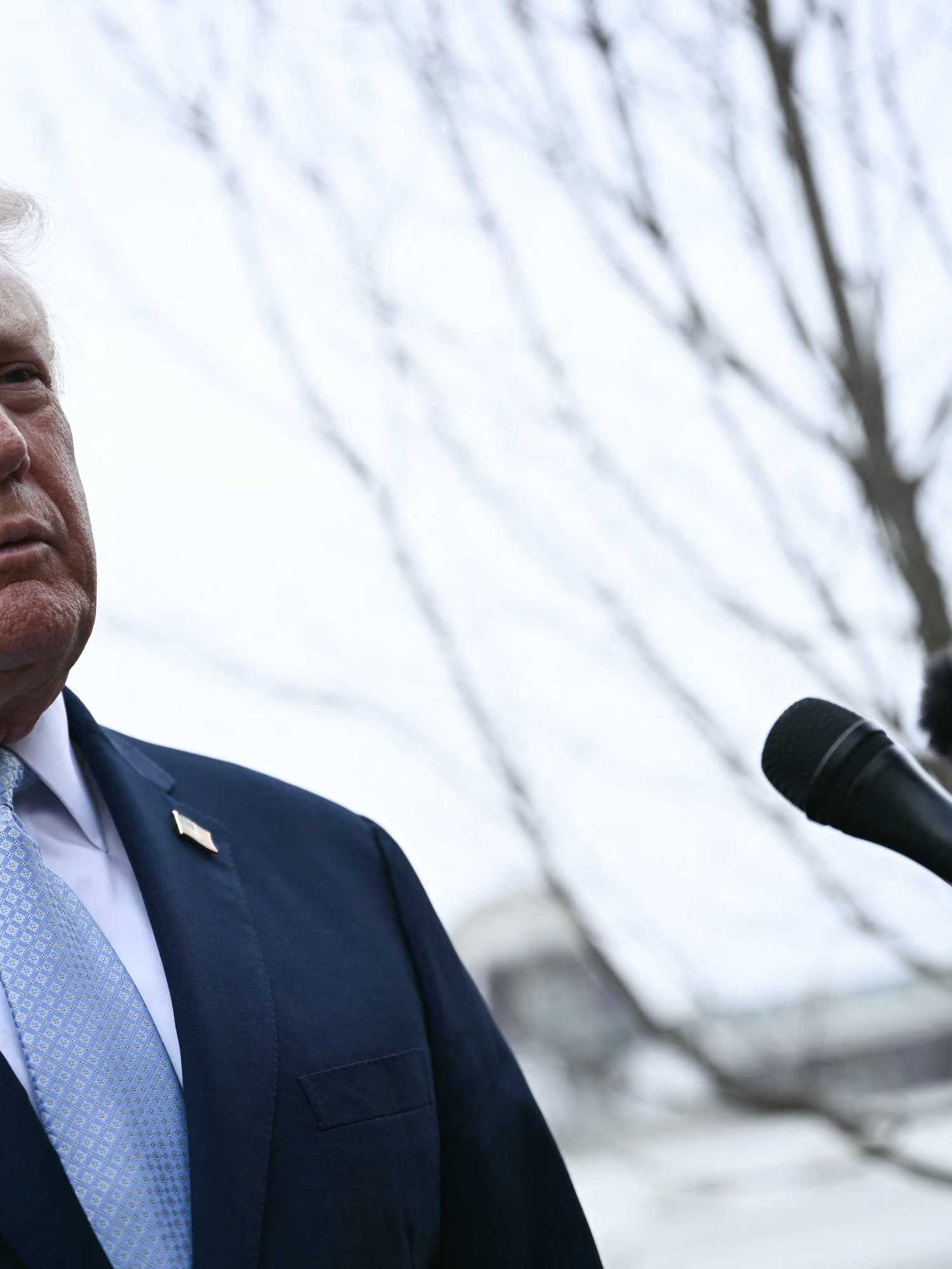 Donald Trump, a white man with short blonde hair in a black suit takes questions from reporters outside the White House.