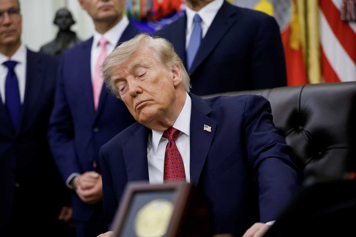 Donald Trump appears to close his eyes while sitting behind the Resolute Desk in the Oval Office. Several men are seen standing behind him.