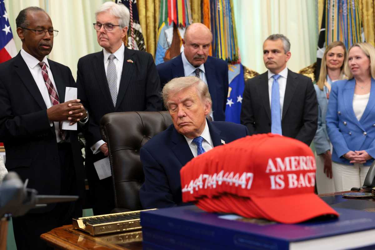Donald Trump closes his eyes while sitting in his chair behind the Resolute Desk in the Oval Office, as a crowd of people stand behind him, with one of them speaking.