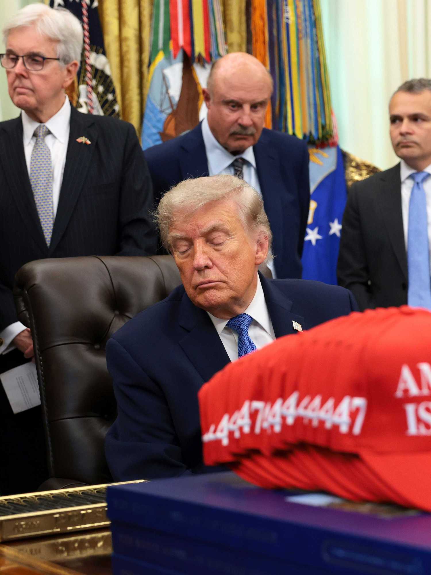 Donald Trump closes his eyes while sitting in his chair behind the Resolute Desk in the Oval Office, as a crowd of people stand behind him, with one of them speaking.