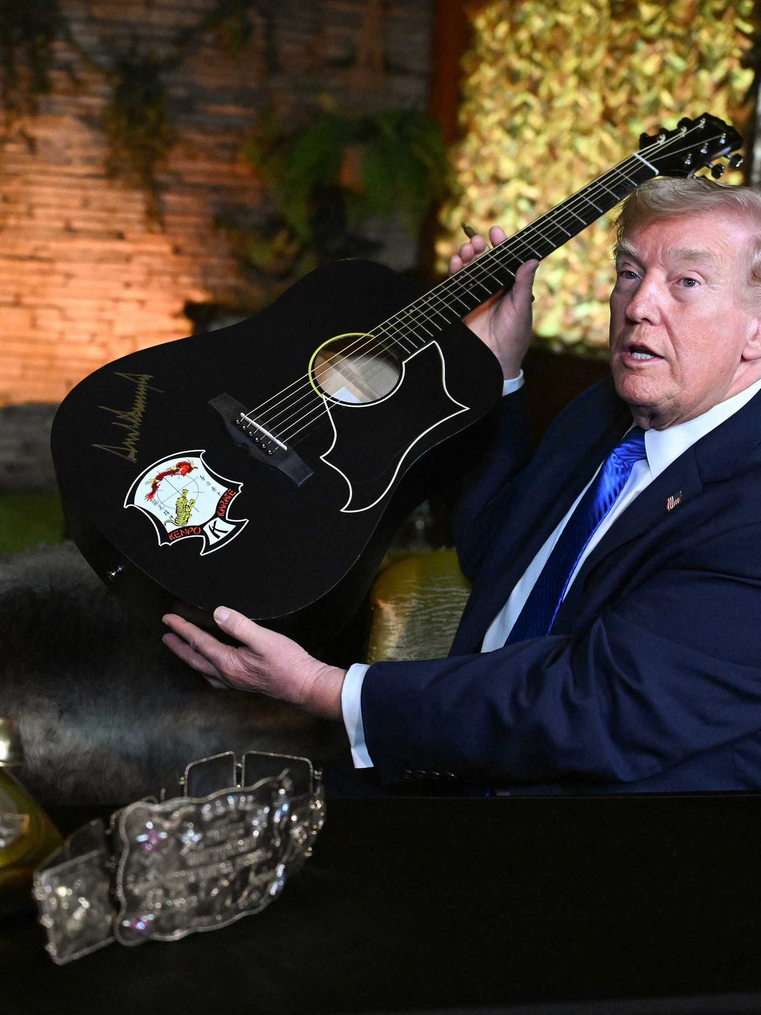 Donald Trump holds up a black acoustic guitar with his autograph on it in golden ink.