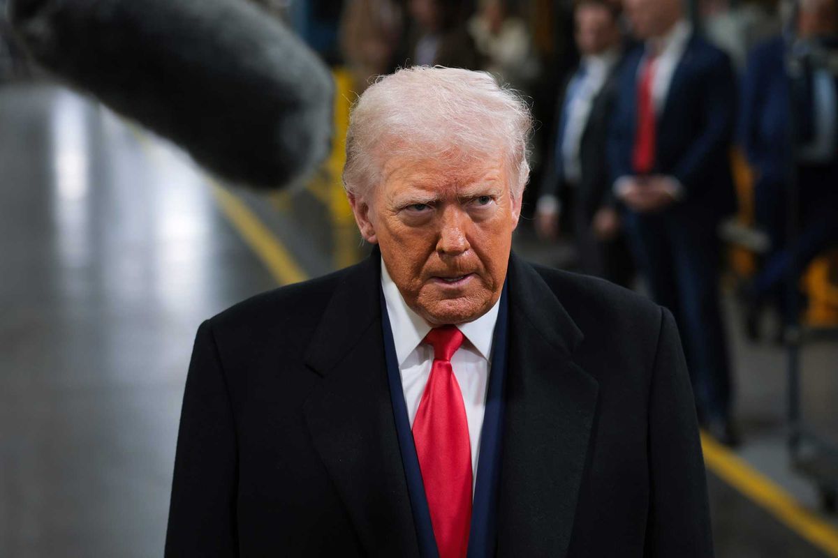 Donald Trump in a black coat, blue suit and red tie, speaking to reporters inside a Ford factory.