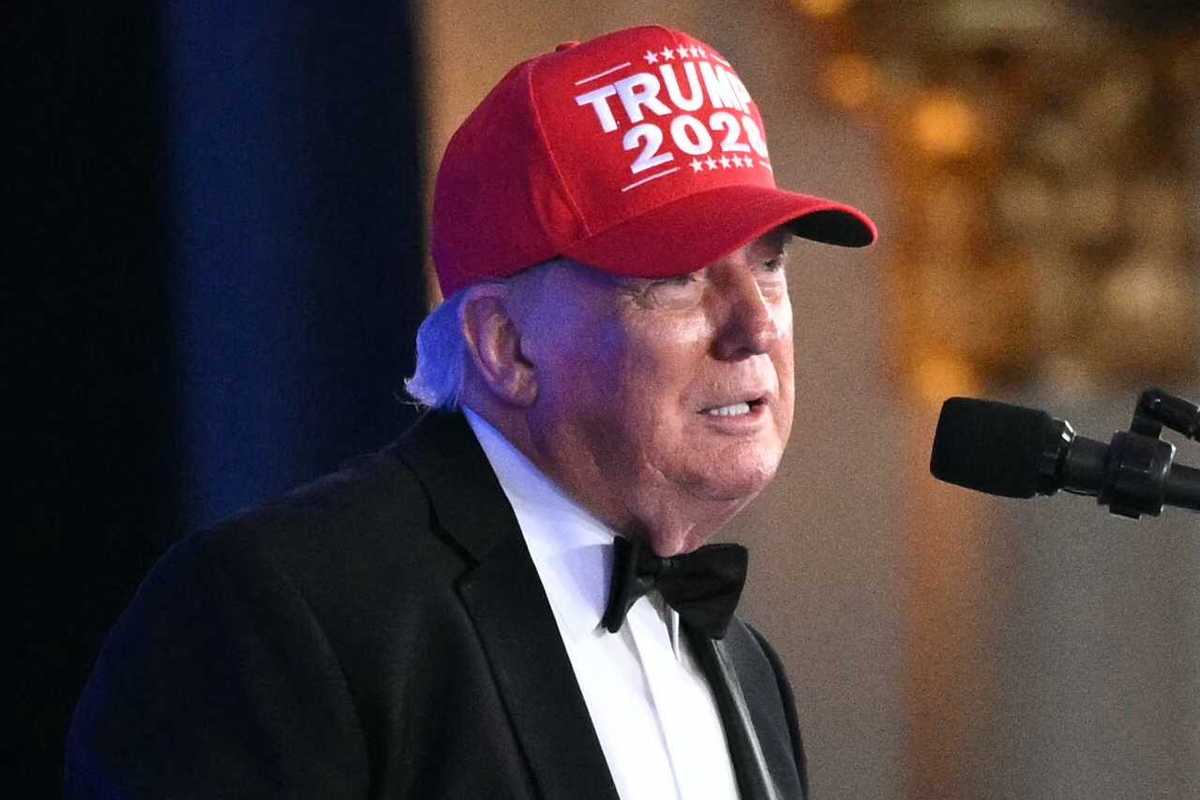 Donald Trump in a black suit and bow tie, with a red 'Trump 2028' hat, speaks into a microphone.