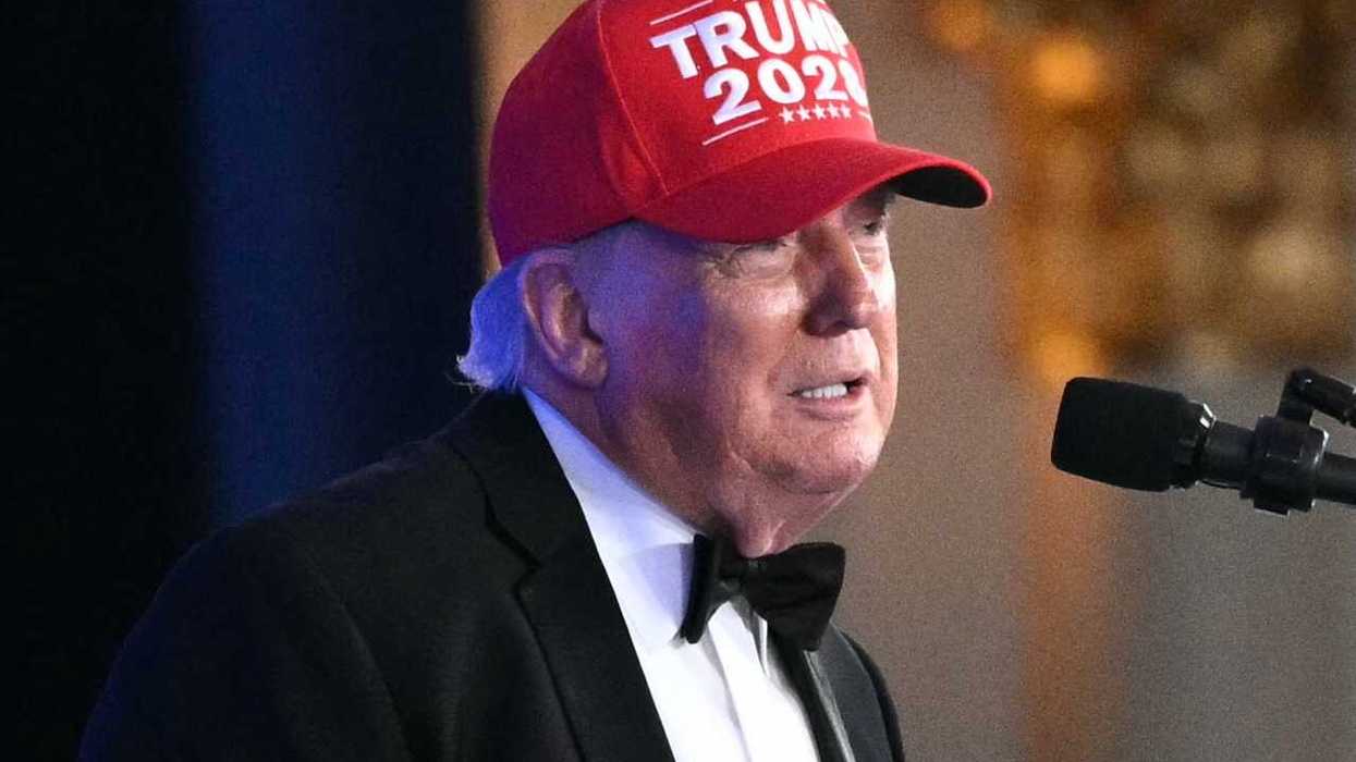 Donald Trump in a black suit and bow tie, with a red 'Trump 2028' hat, speaks into a microphone.