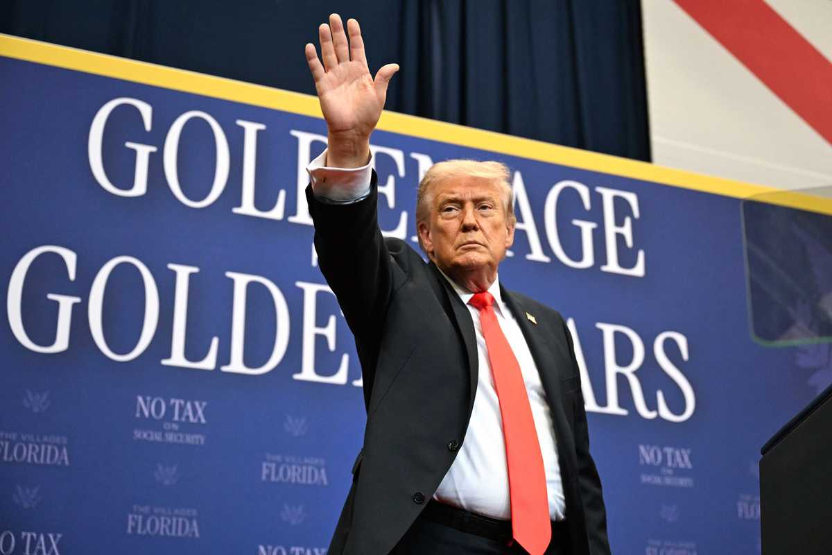 Donald Trump in a black suit and red tie waves at a crowd while standing in front of a blue poster board with the words 'Golden age, golden years'.