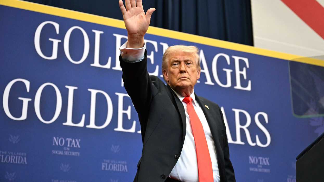 Donald Trump in a black suit and red tie waves at a crowd while standing in front of a blue poster board with the words 'Golden age, golden years'.