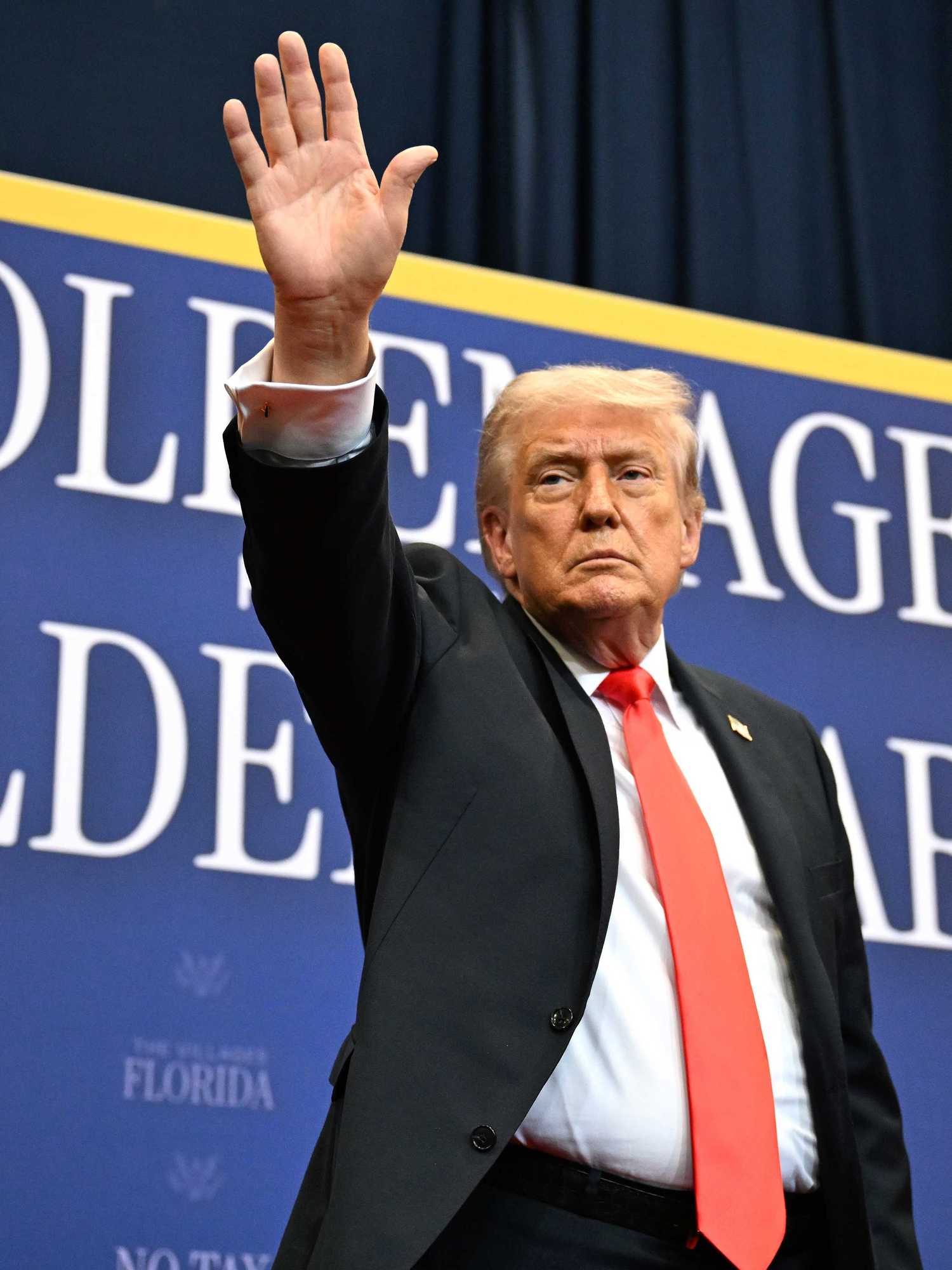 Donald Trump in a black suit and red tie waves at a crowd while standing in front of a blue poster board with the words 'Golden age, golden years'.