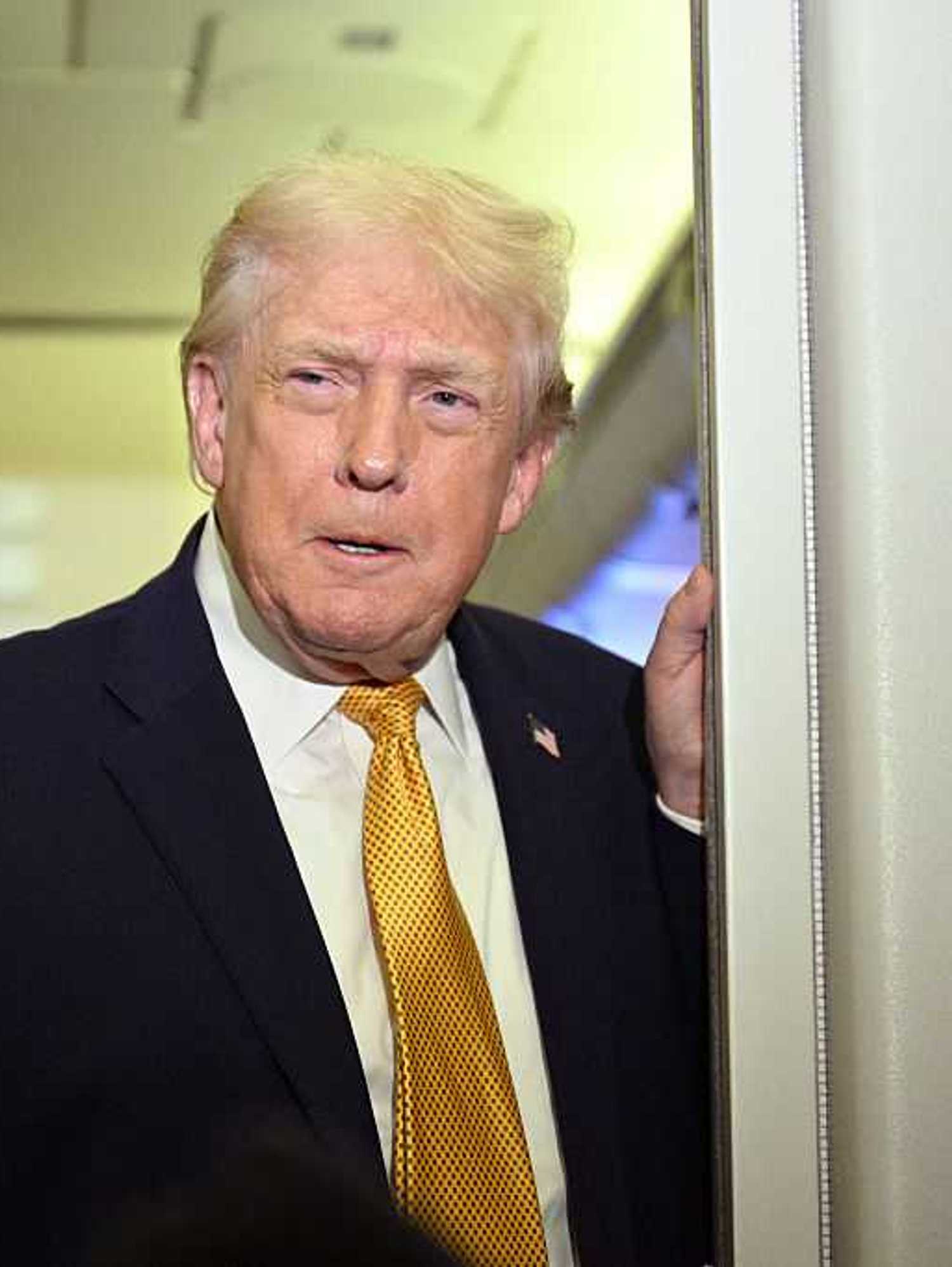 Donald Trump in a black suit and yellow tie talks to reporters on board Air Force One.