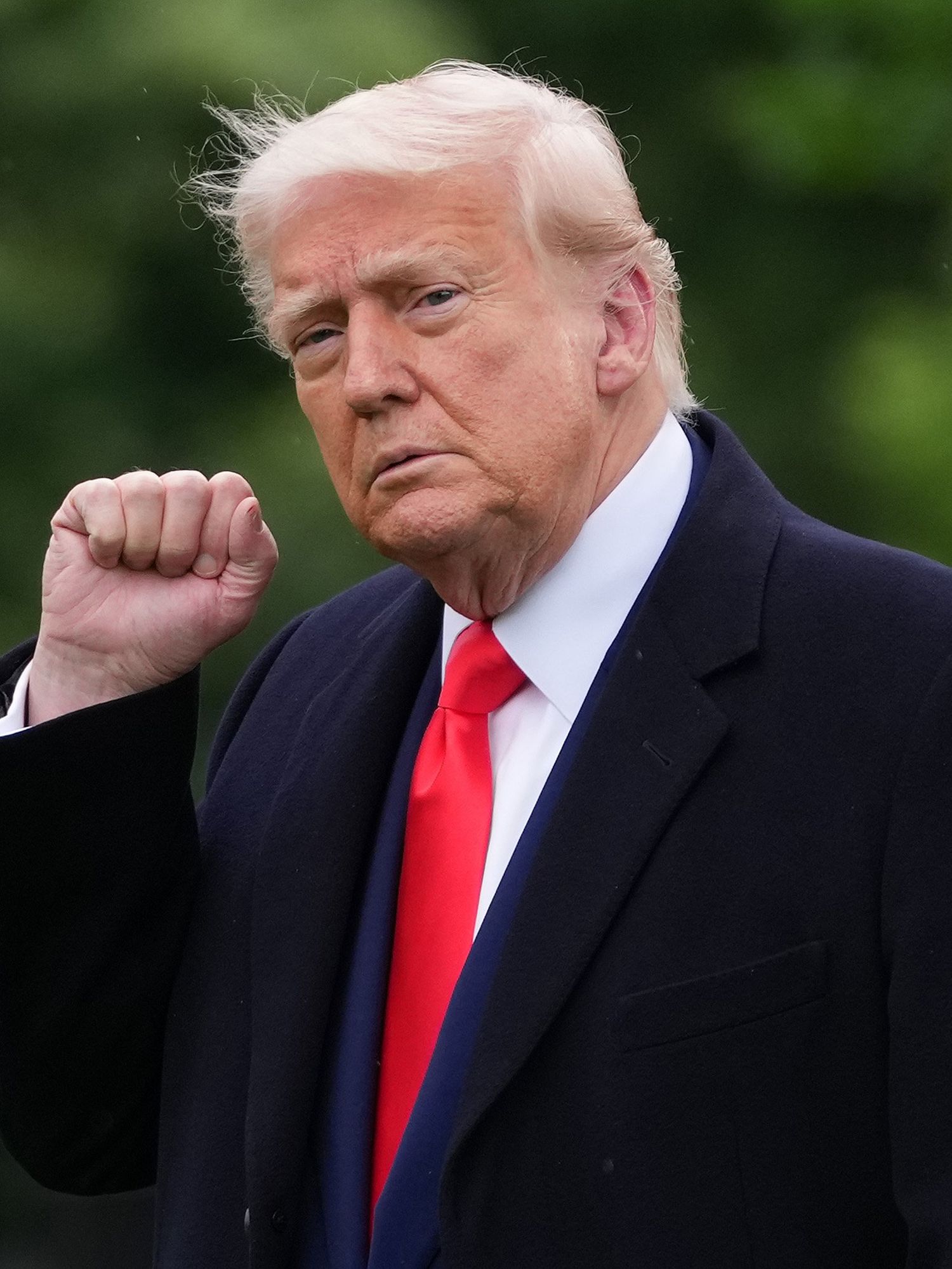 Donald Trump, in a black suit jacket, blue suit and red tie, raises his right fist in the air with a neutral expression on his face as he departs the White House.