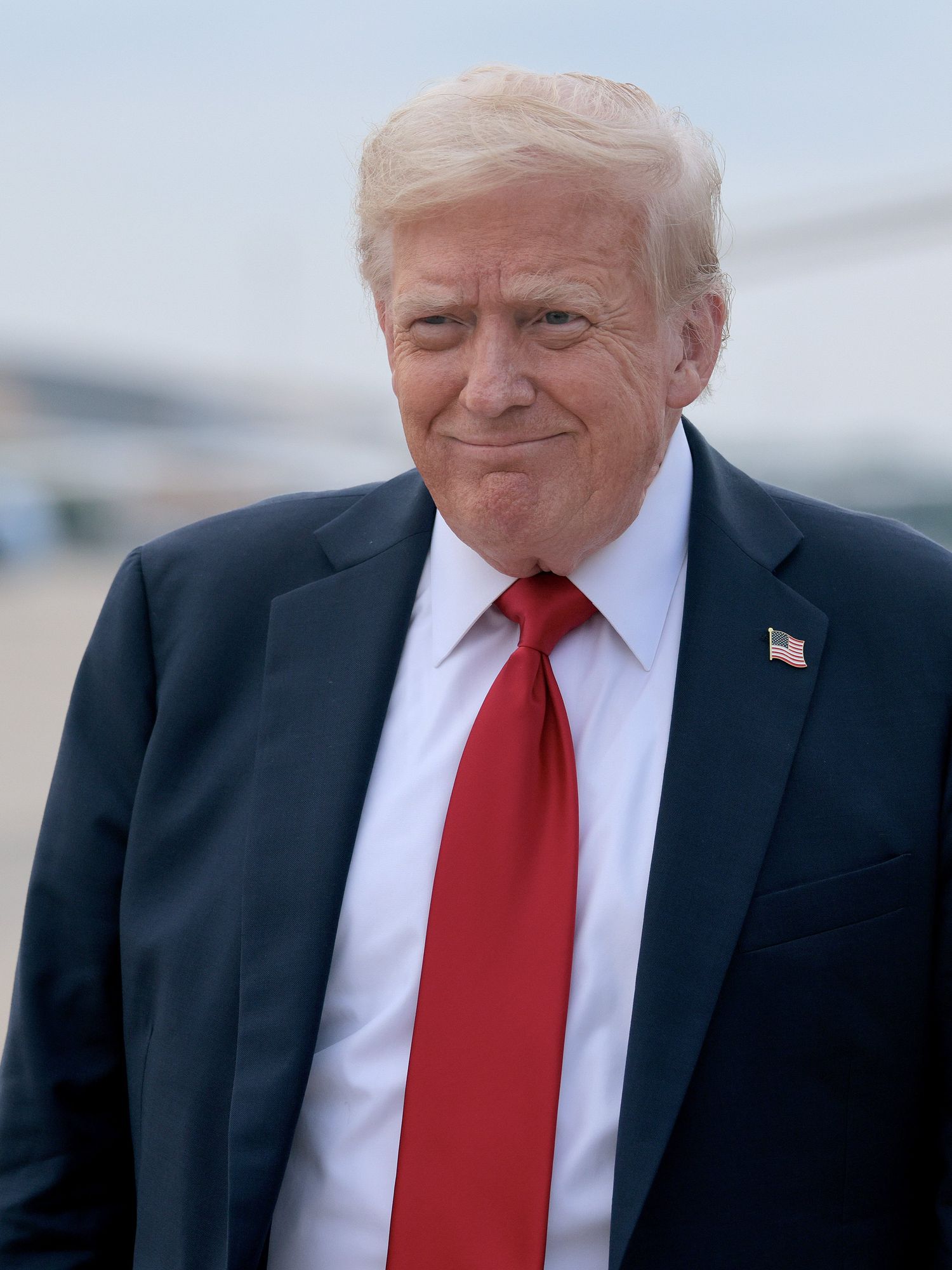 Donald Trump, in a black suit with a red tie, smiles as he stands on an airfield.