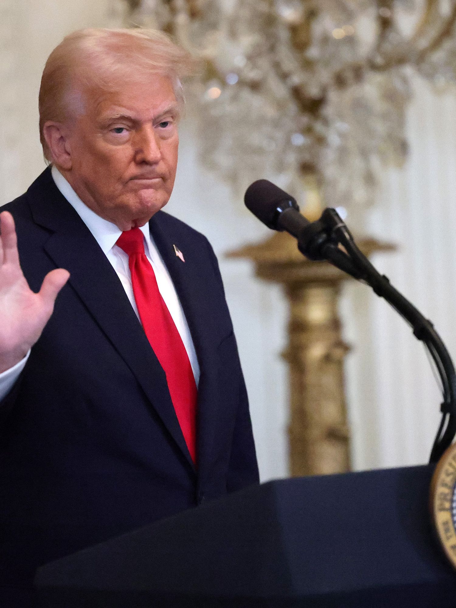 Donald Trump, in a black suit with a red tie, stands behind his presidential lectern in the White House and raises his right hand to wave, with a neutral expression on his face.