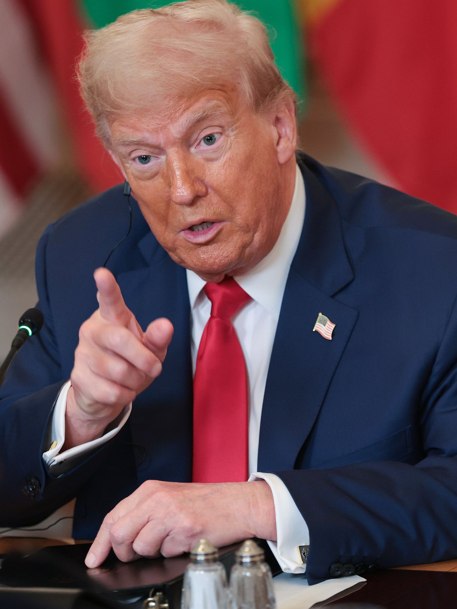 Donald Trump in a blue suit and red tie speaks behind a desk, pointing with his right index finger.