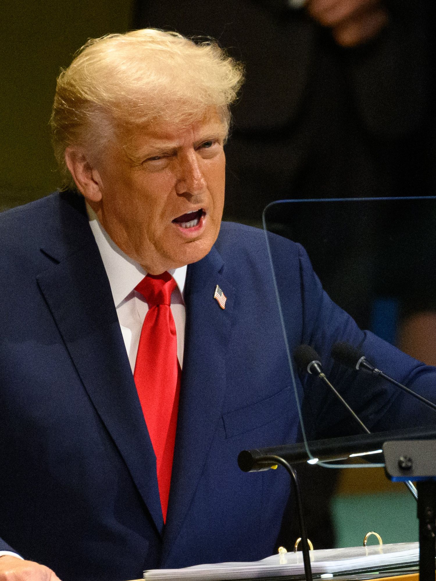 Donald Trump in a blue suit and red tie speaks behind a wooden lectern at the UN General Assembly.