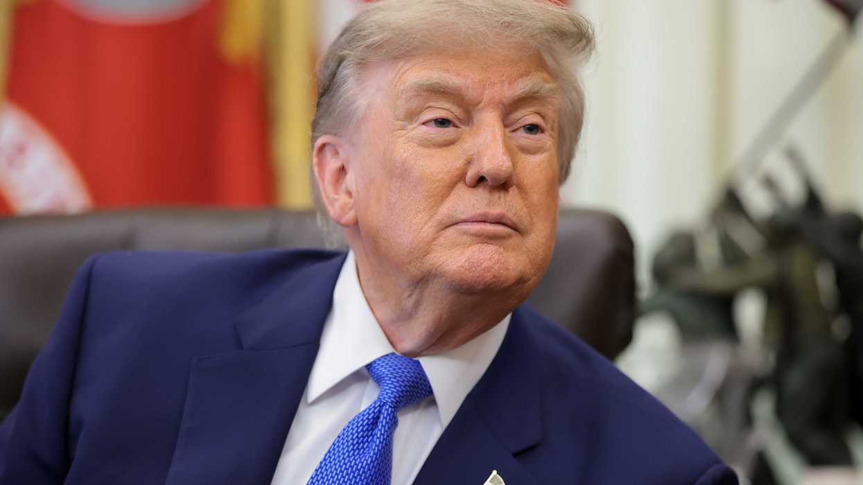 Donald Trump, in a blue suit and tie, looks at someone off-camera in the Oval Office. He's sat behind the Resolute Desk.