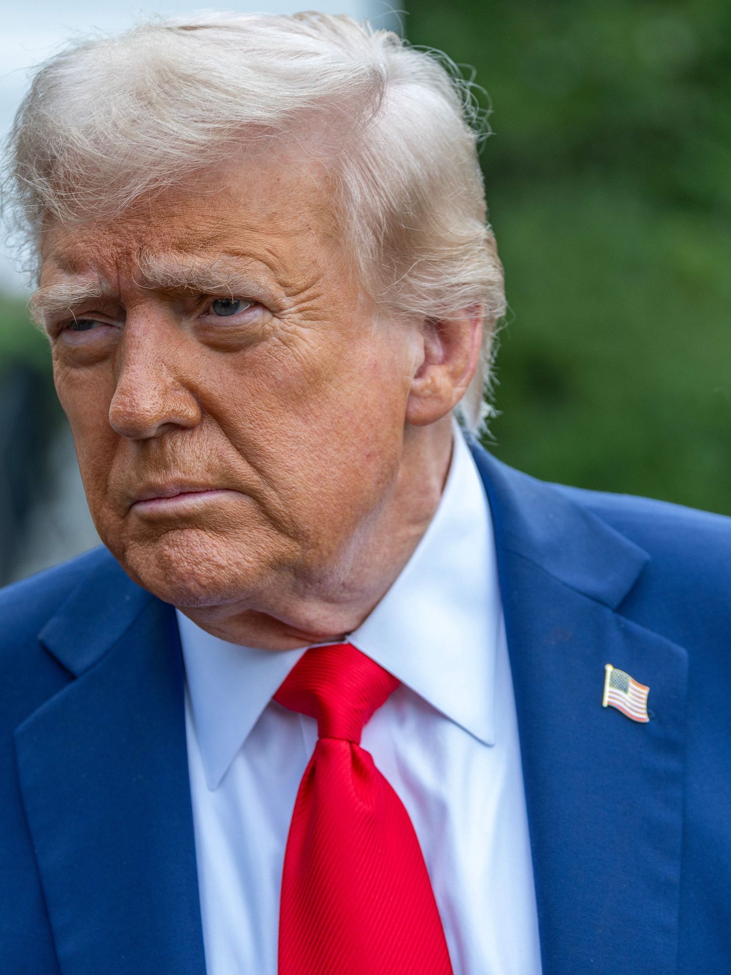 Donald Trump in a blue suit outside the White House, looking to his right with a neutral expression on his face.