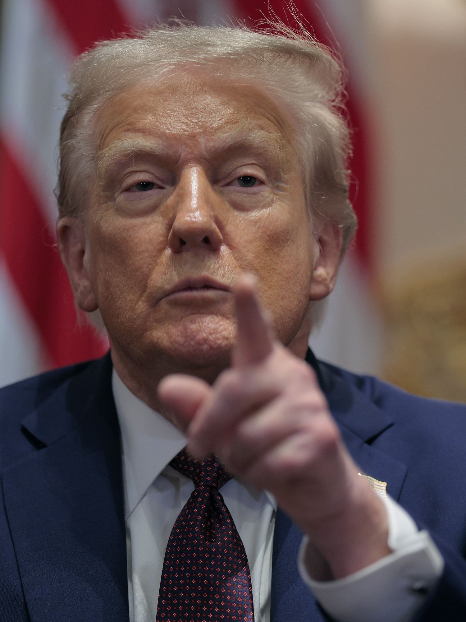 Donald Trump in a blue suit points his left index finger at the camera with a neutral expression on his face.