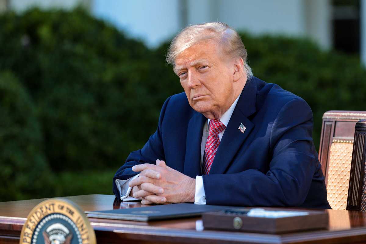 Donald Trump in a blue suit sitting outside the White House behind a desk with the presidential seal on it.