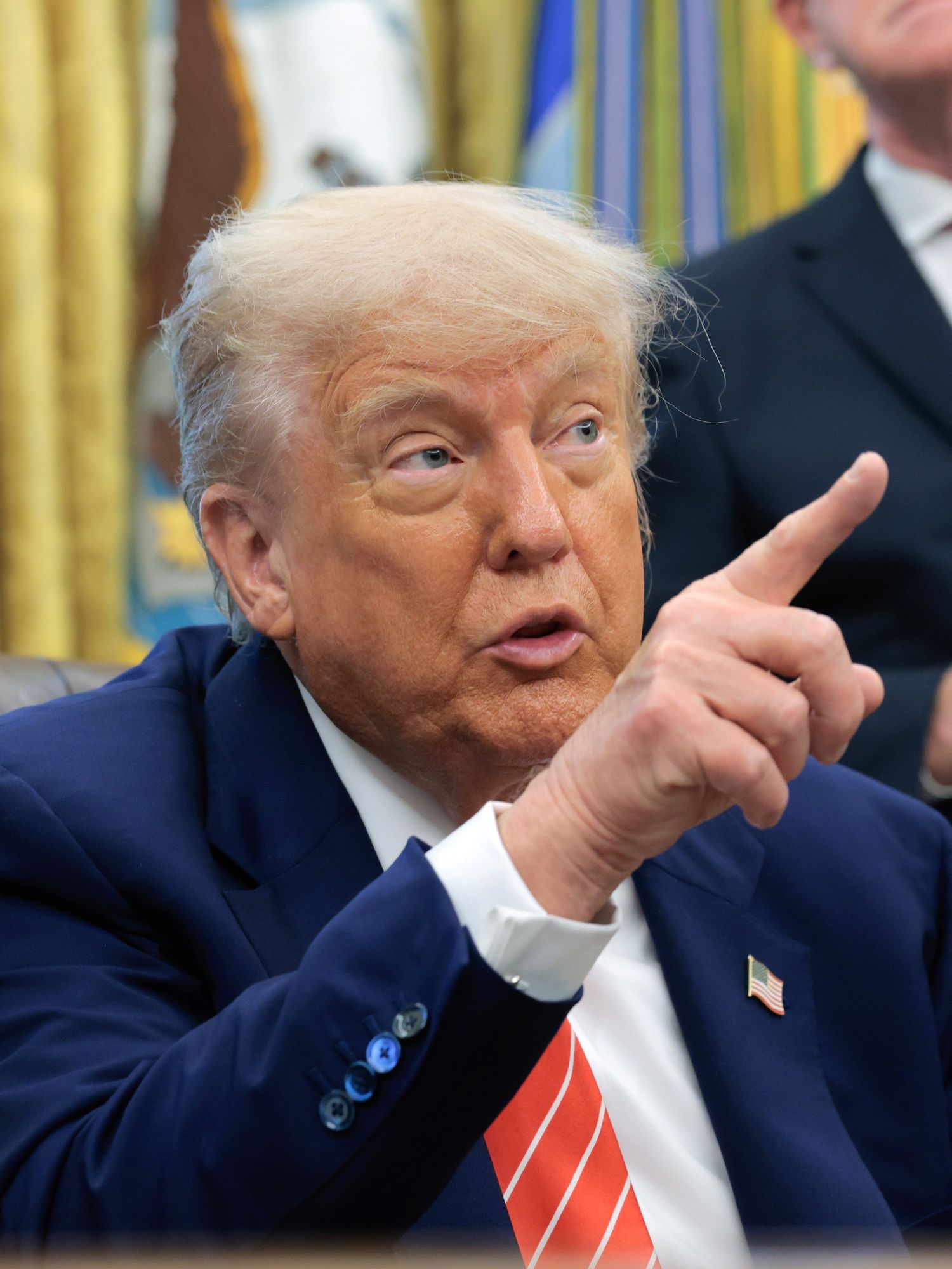 Donald Trump, in a blue suit with a striped red tie, sits behind his desk in the Oval Office, pointing at something with his right index finger.