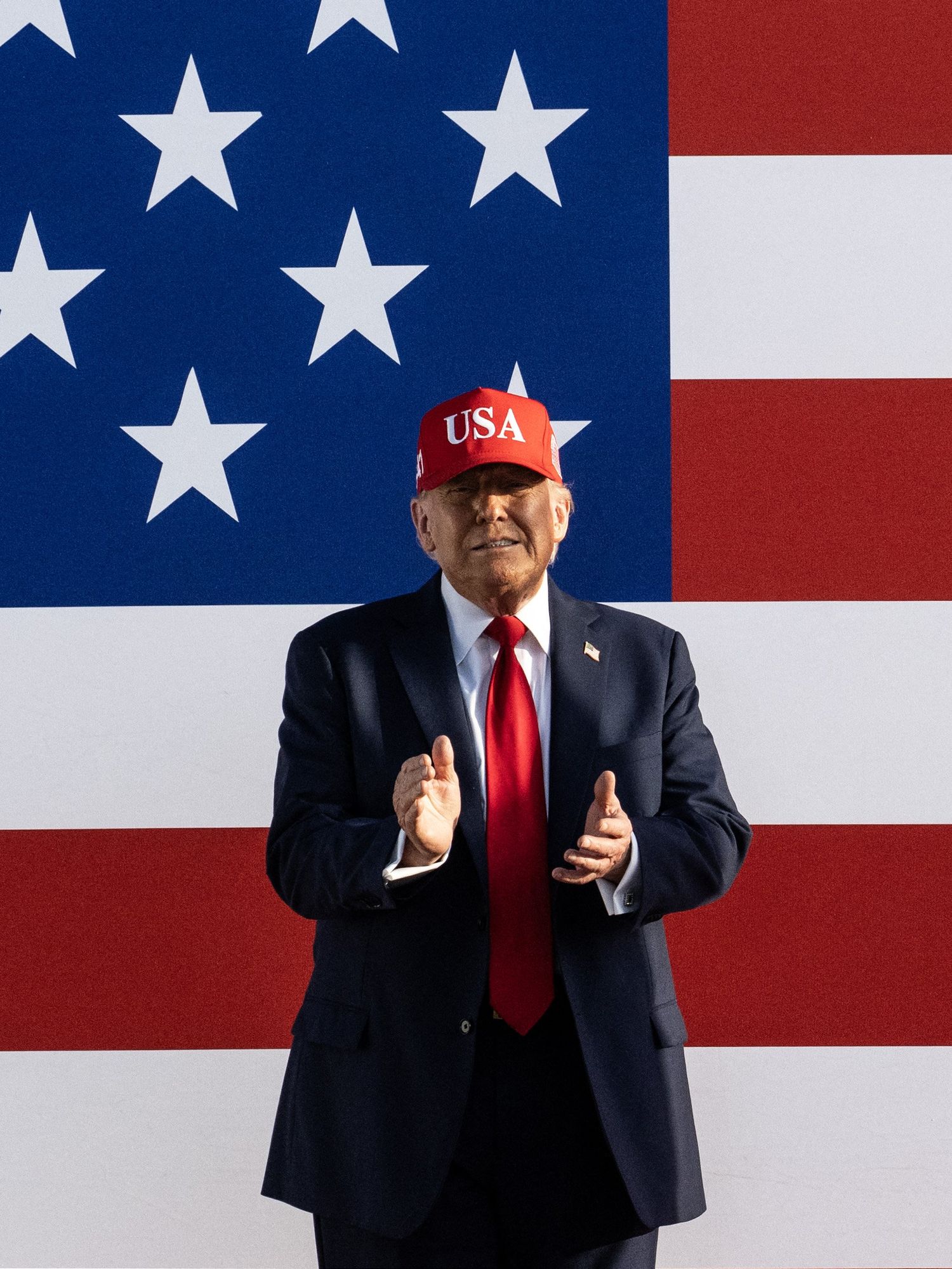 Donald Trump, in a dark black suit and red USA cap, smiles and claps behind a giant American flag.